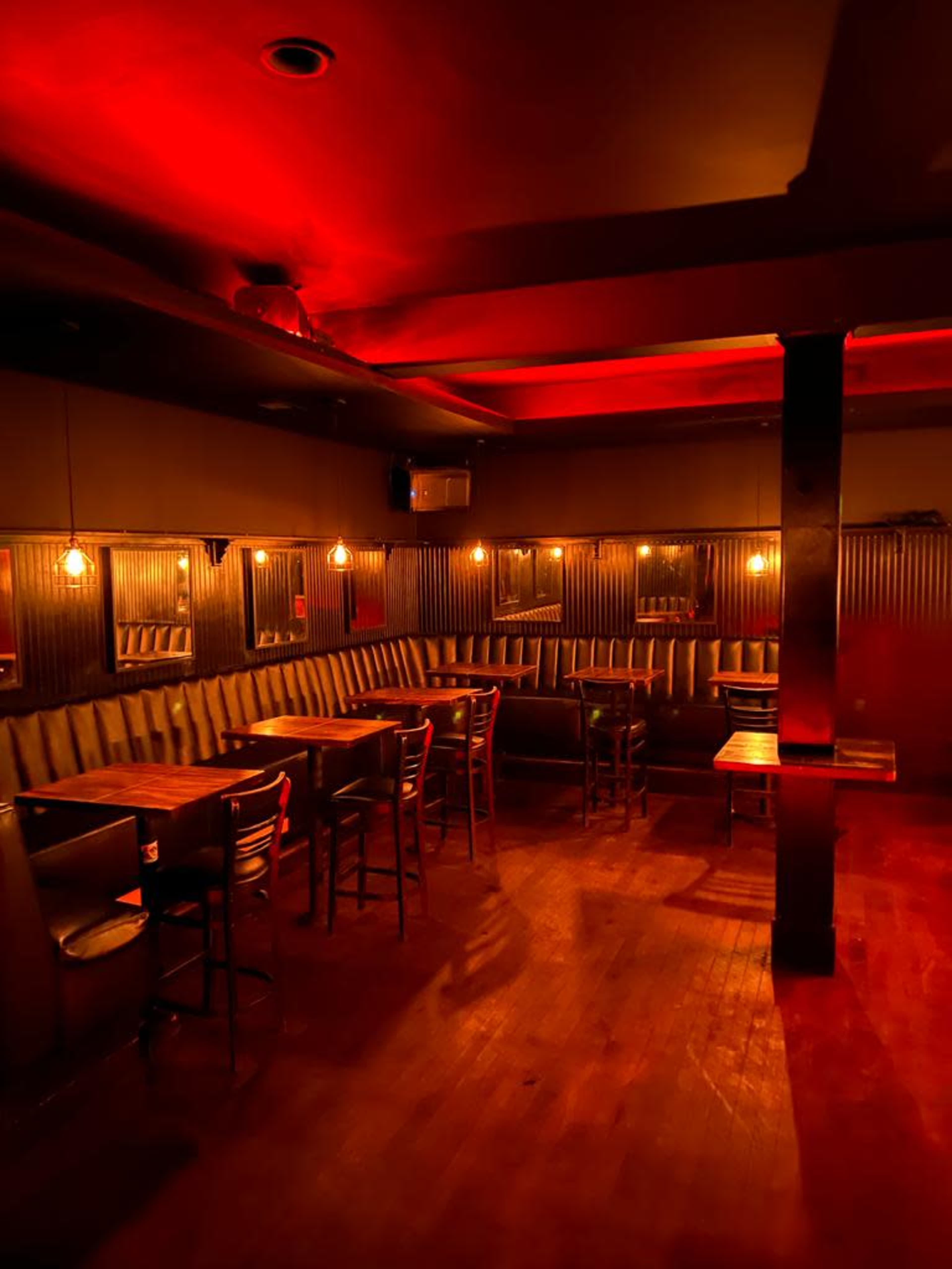A dimly lit bar with a wooden floor, metal-paneled walls, and several empty tables and chairs.
