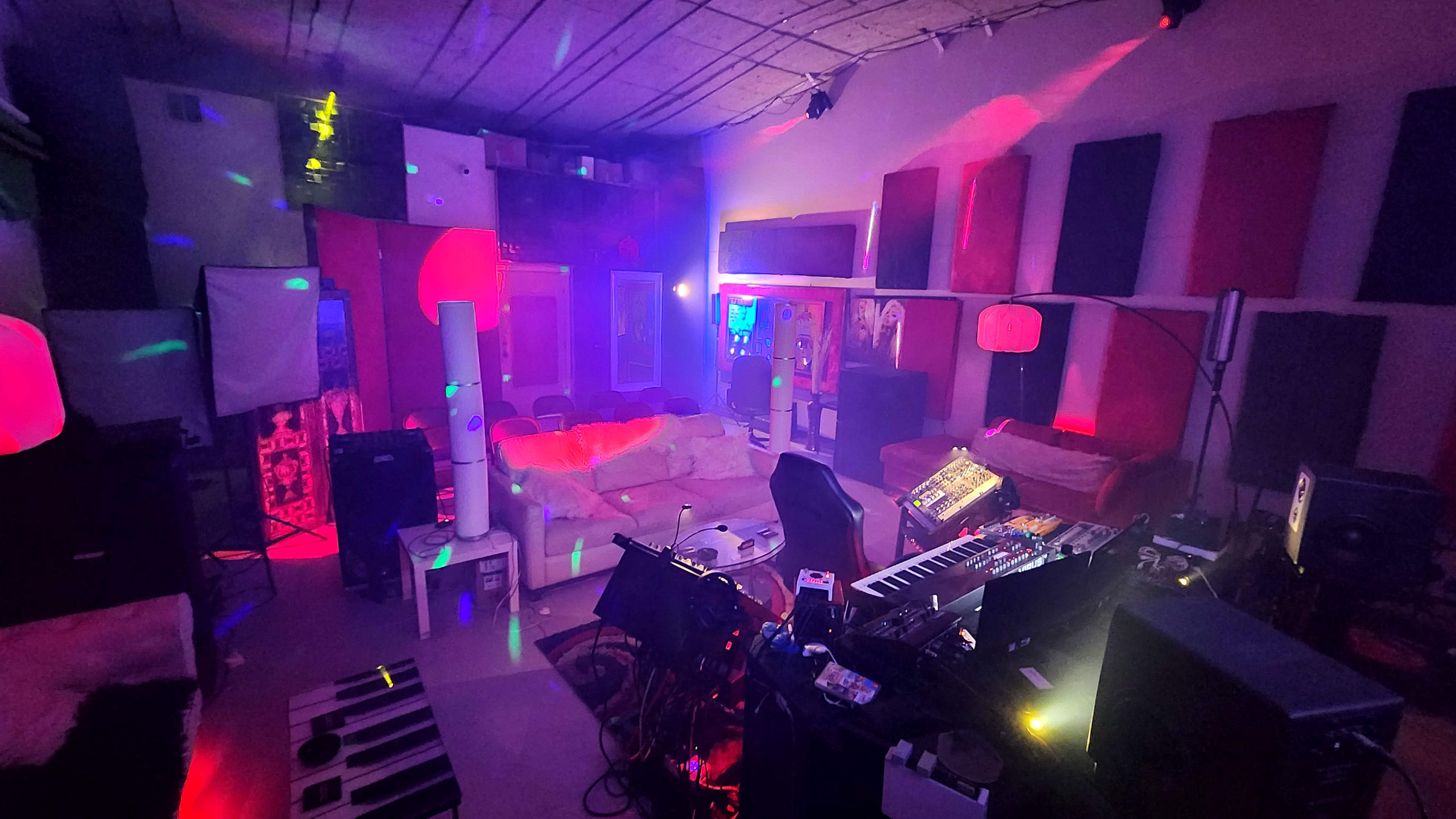 A music studio with various synthesisers, keyboards, and seating areas, illuminated by colorful lights and soundproofing panels on the walls.