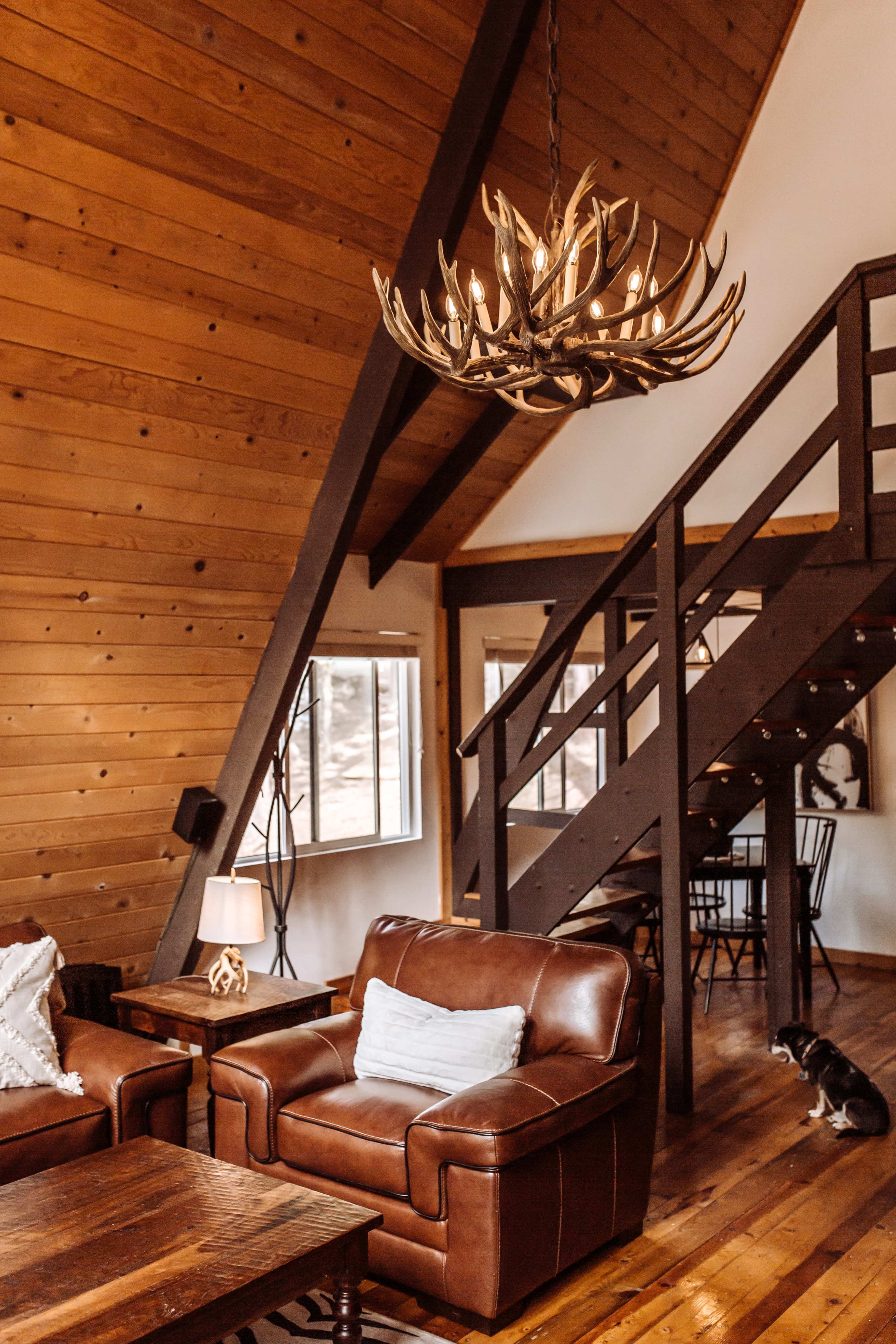 A rustic living room with a leather couch, wooden flooring, and a large chandelier made of antlers, alongside a staircase leading to the upper level.