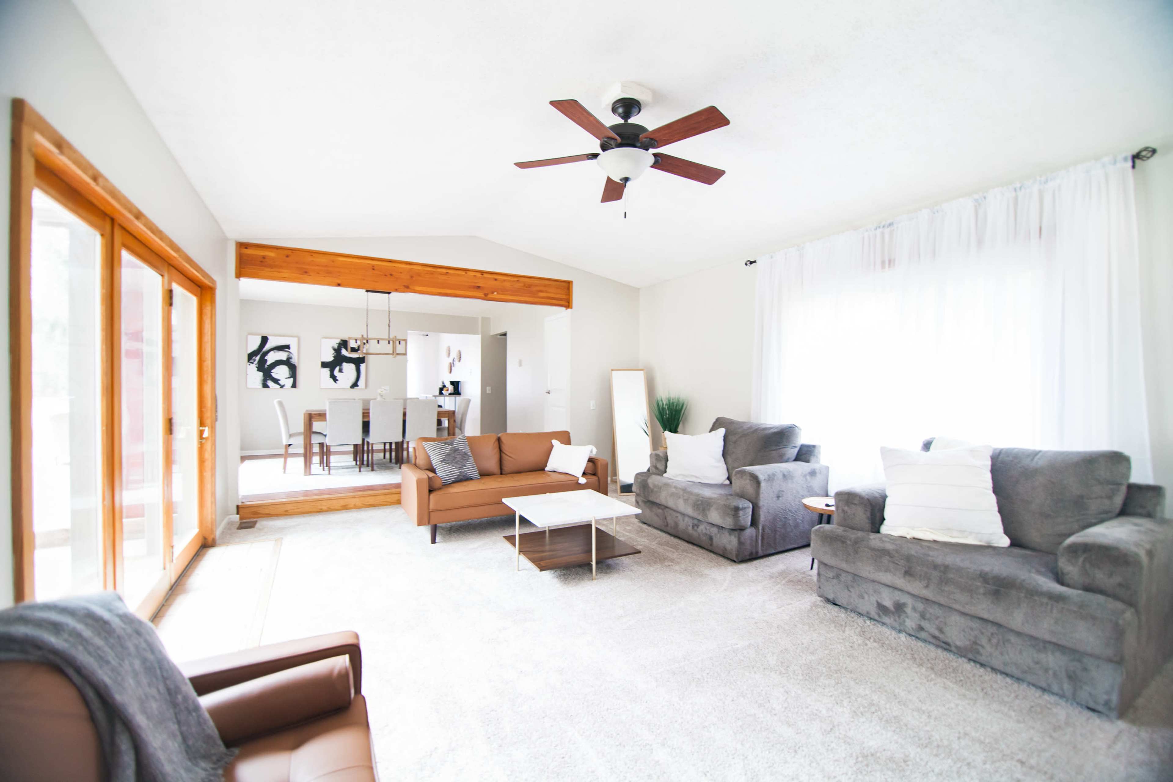 The living room features a light-colored carpet, a ceiling fan, and seating arranged around a coffee table, with a dining area visible in the background.