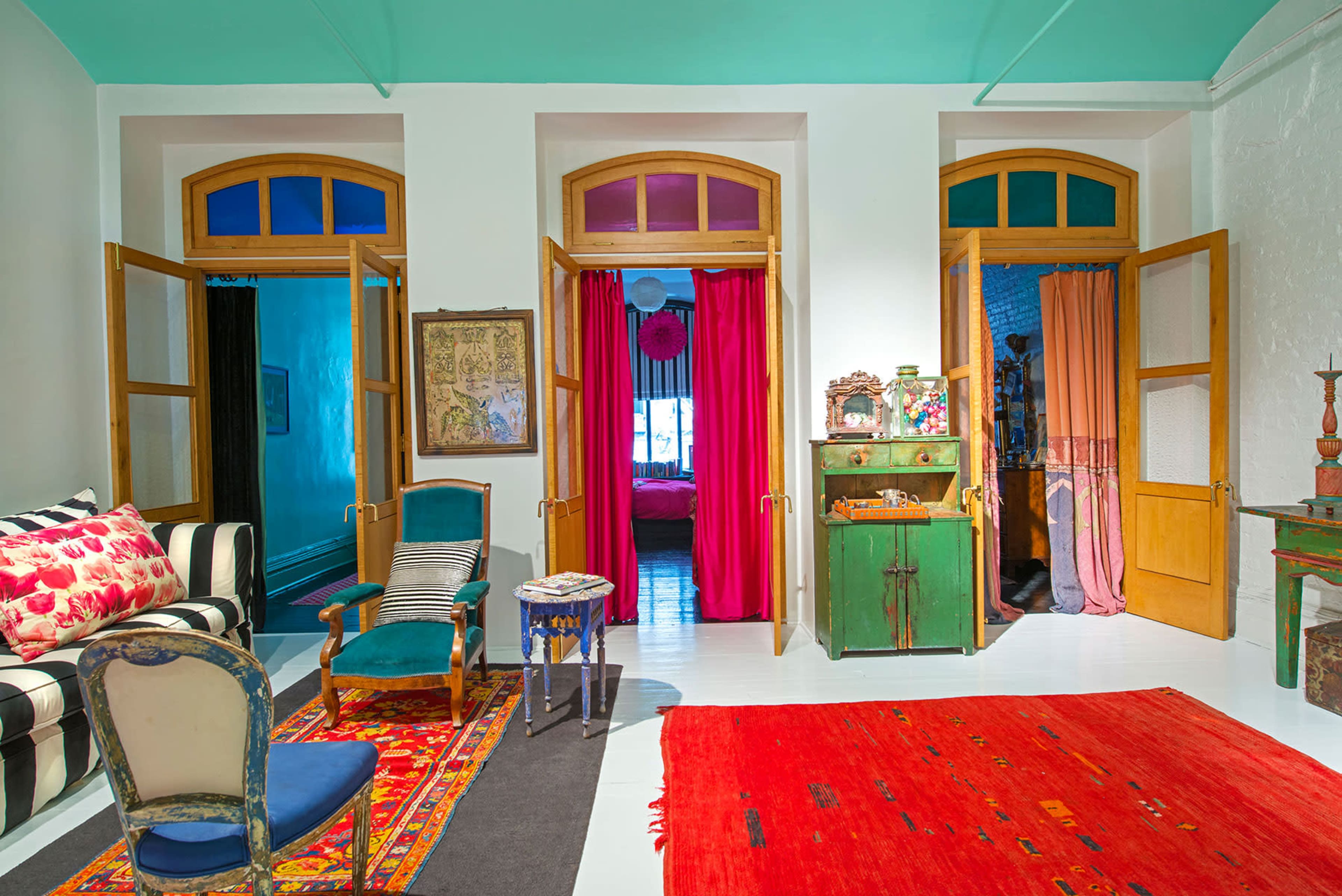 A brightly colored room features three doorways with open doors, leading to adjacent spaces, and is decorated with a variety of furniture and rugs.