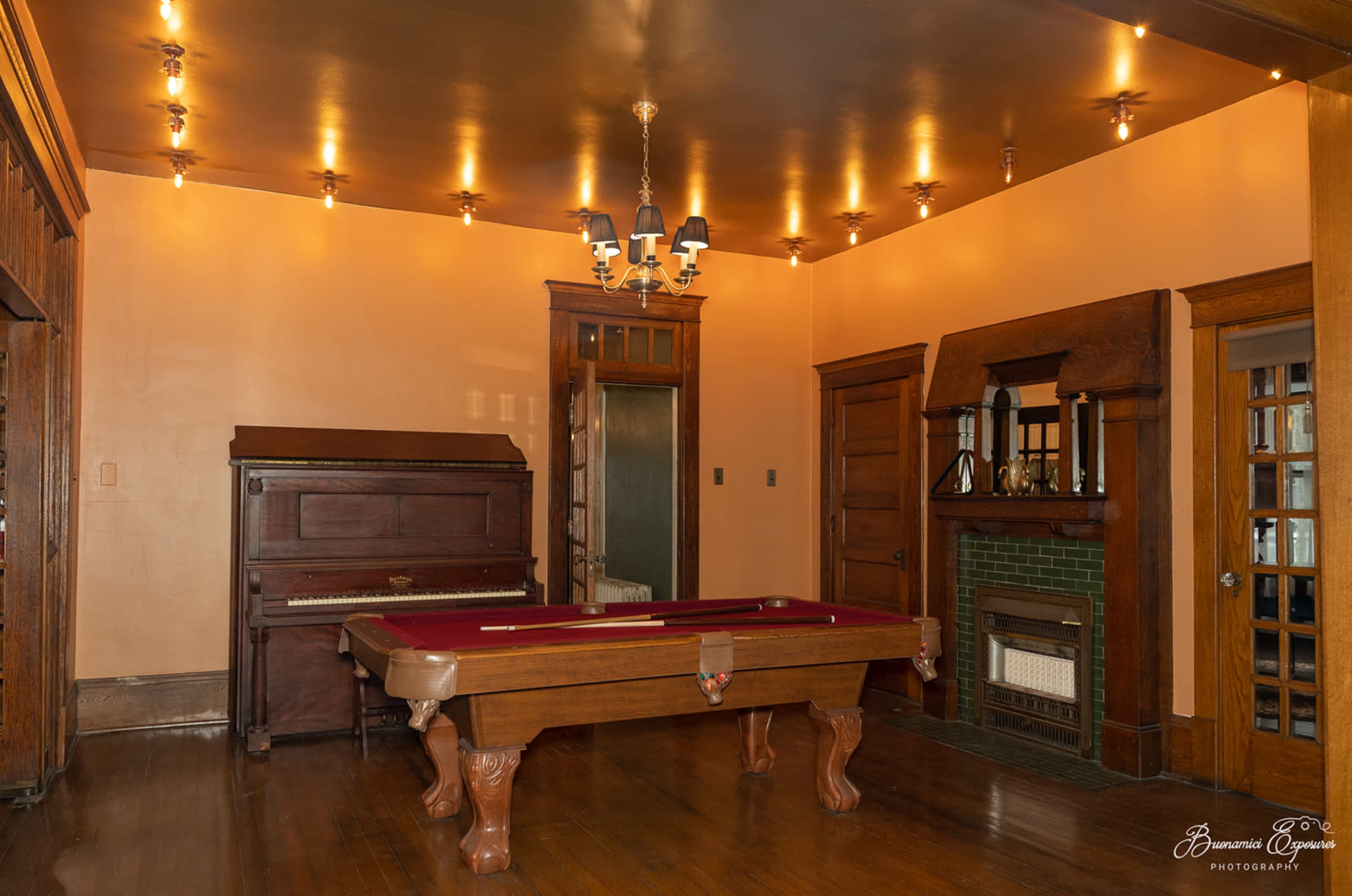 The image shows an interior room featuring a billiard table, a vintage piano, and a fireplace surrounded by wooden paneling.