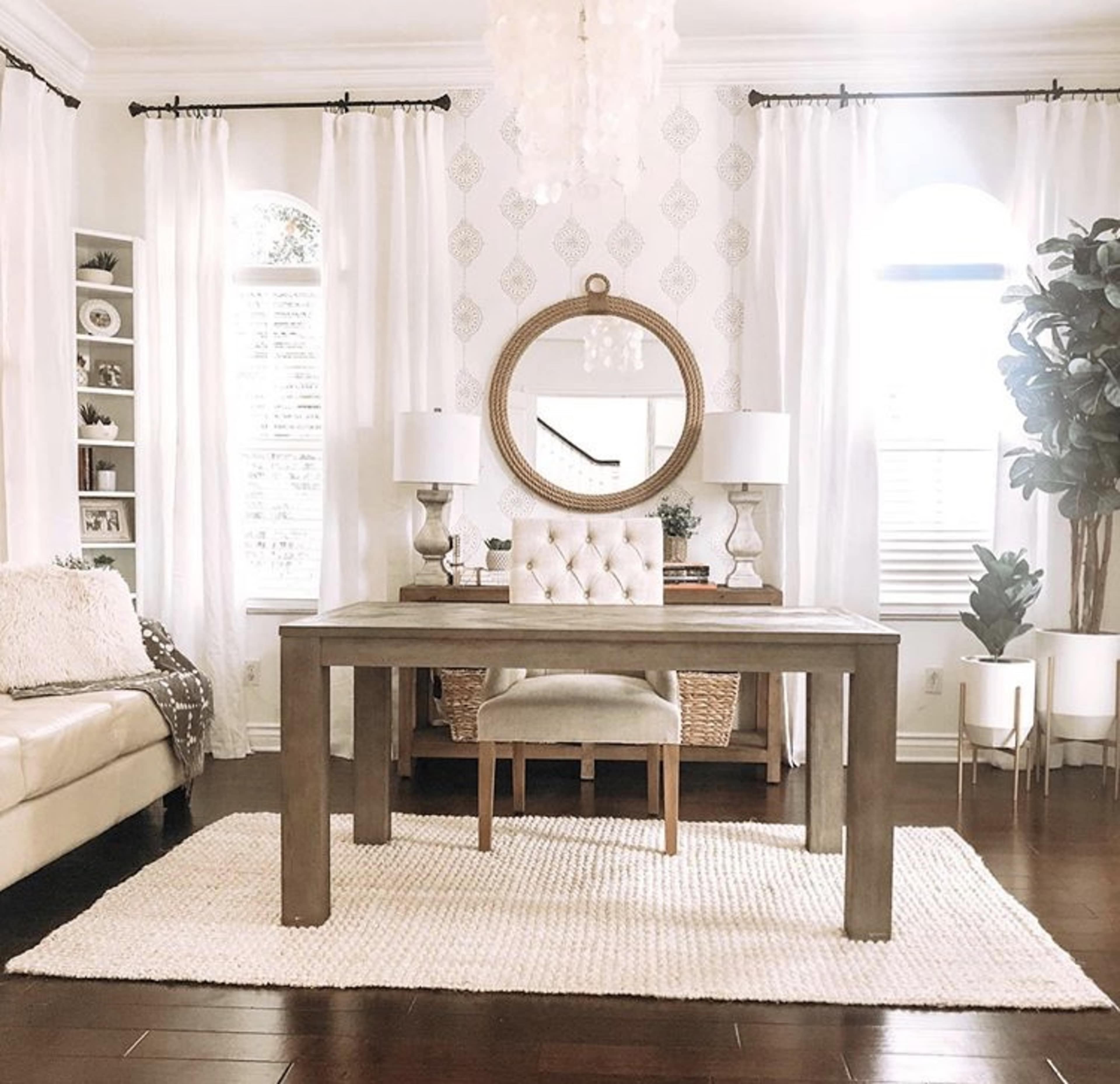 A modern home office with a large table, an upholstered chair, and decor including a round mirror and two lamps, all set against light-colored curtains and a patterned wall.