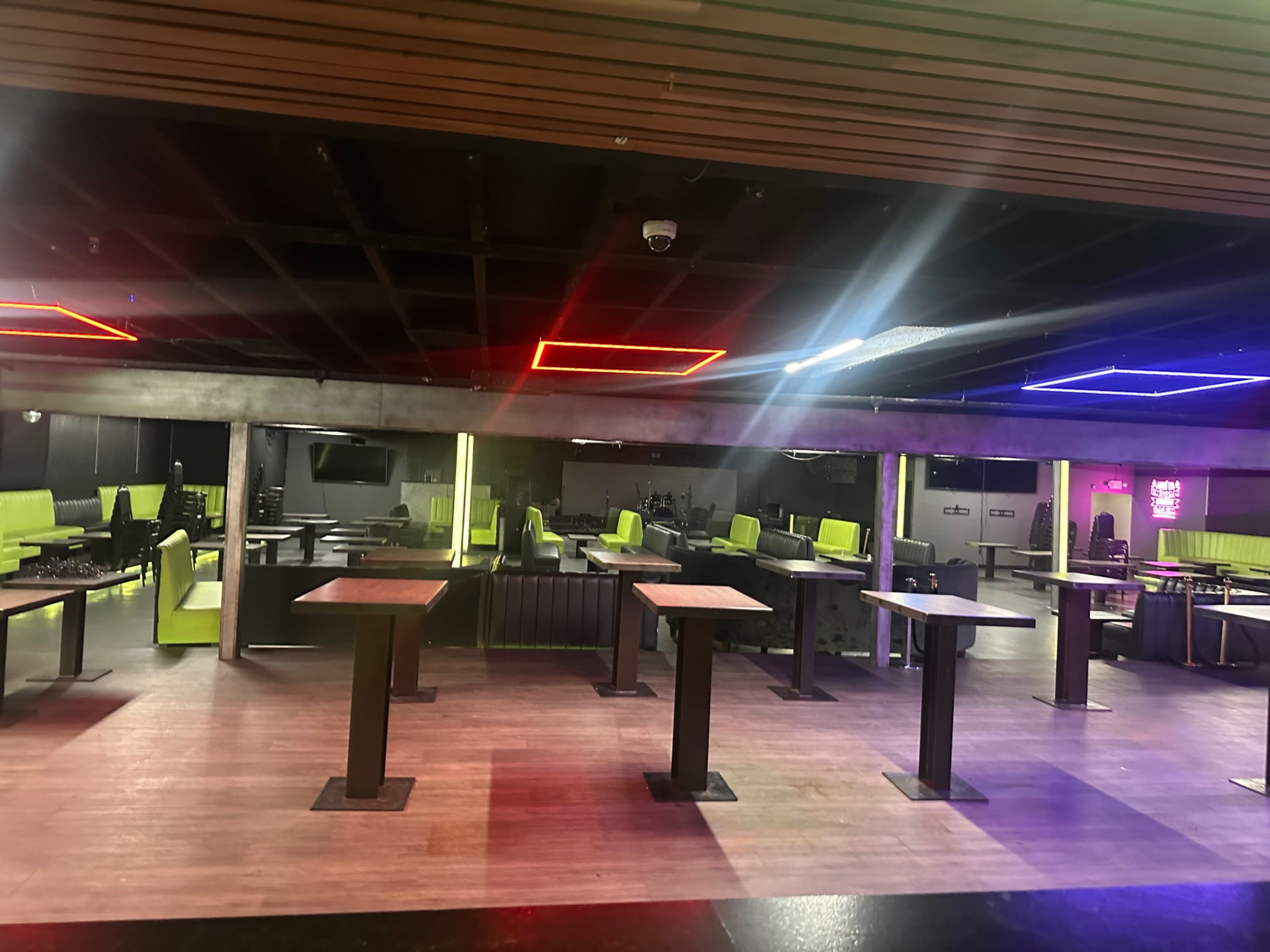 The image shows an empty dining area with green chairs and tables, illuminated by colorful neon lights on the ceiling.