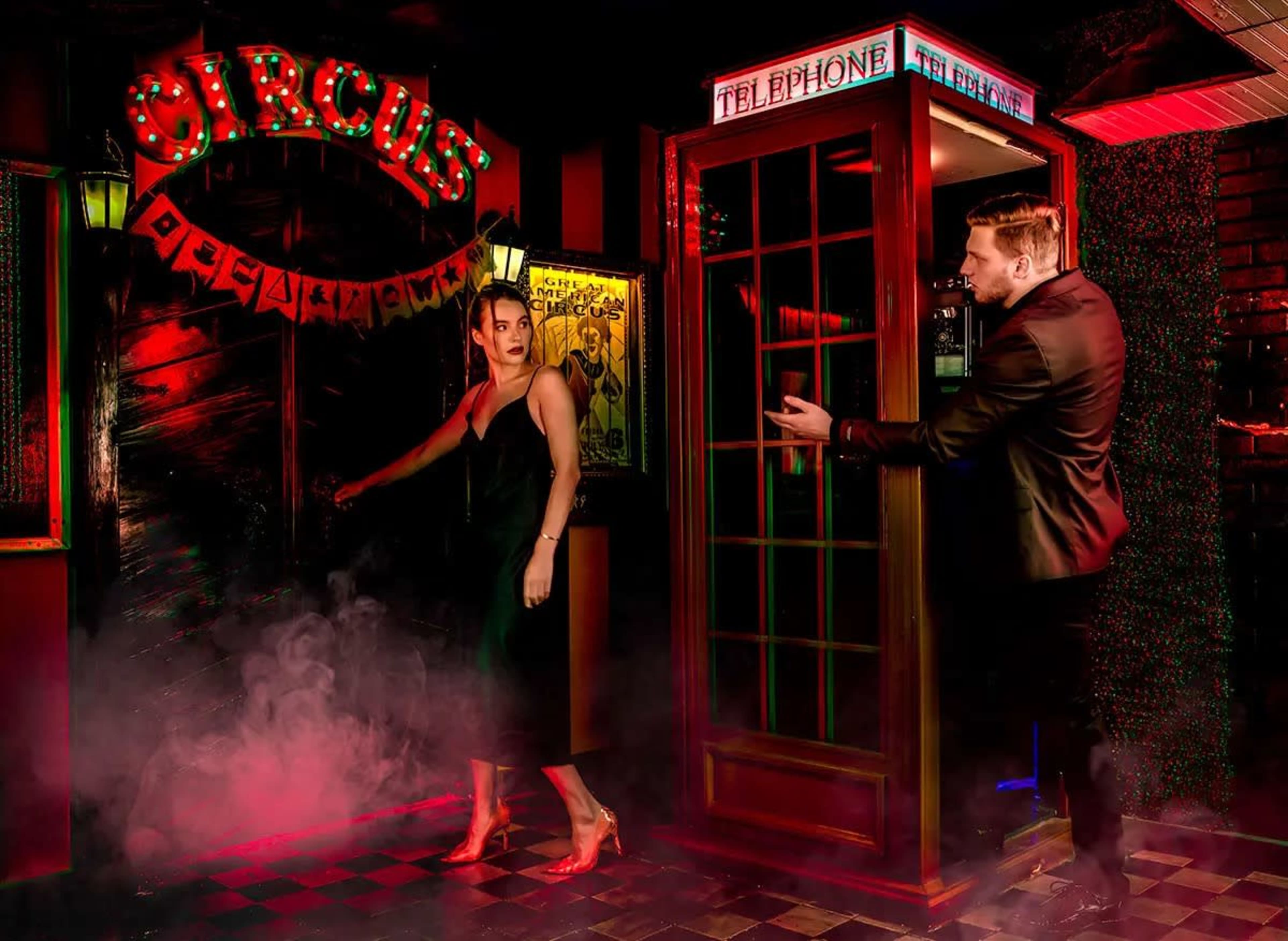 A woman in a black dress exits a bar illuminated by neon signs, while a man in a suit gestures from a nearby telephone booth.