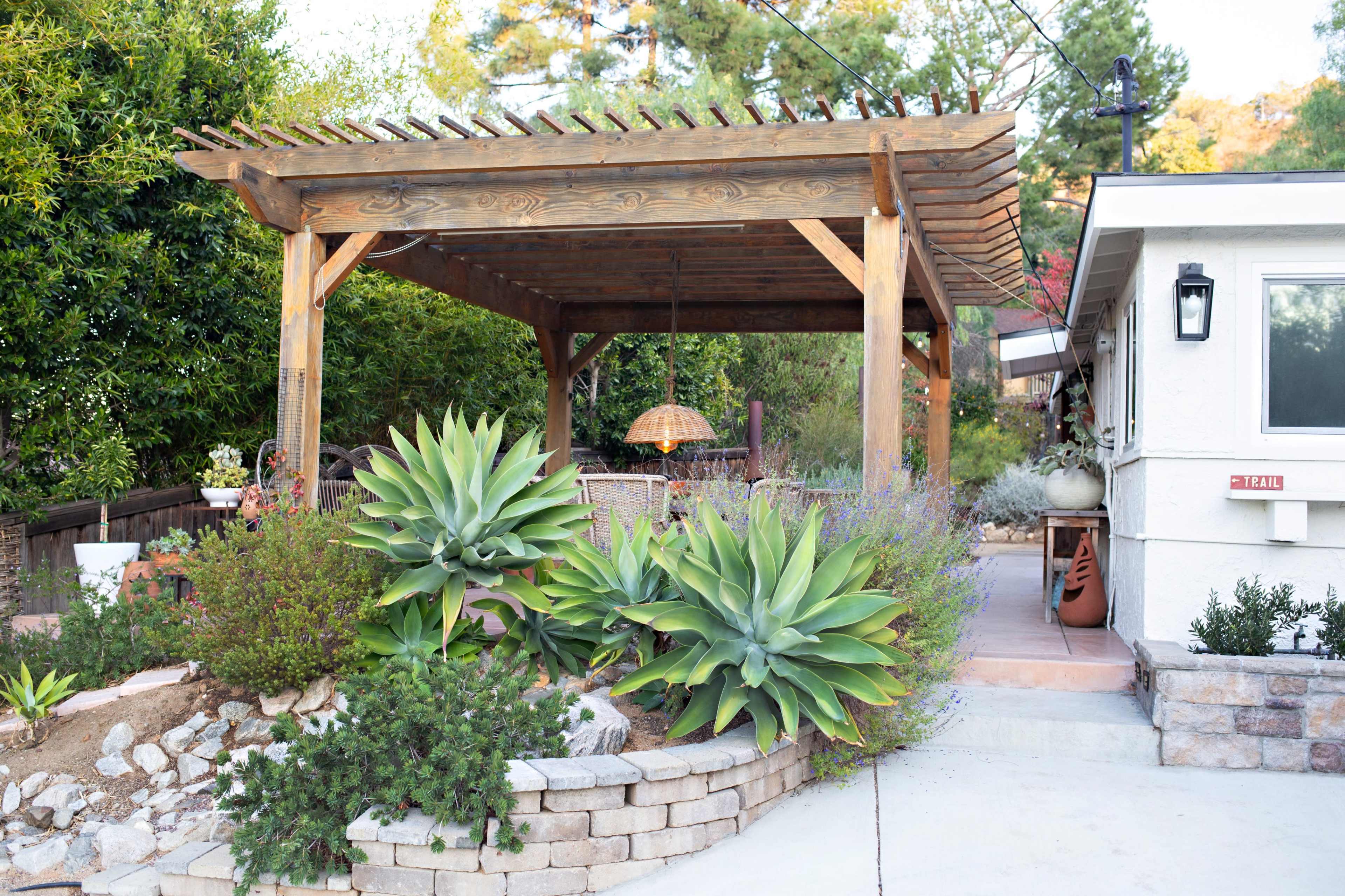 A wooden pergola with a hanging light fixture overlooks a landscaped garden featuring large succulents and a patio area.