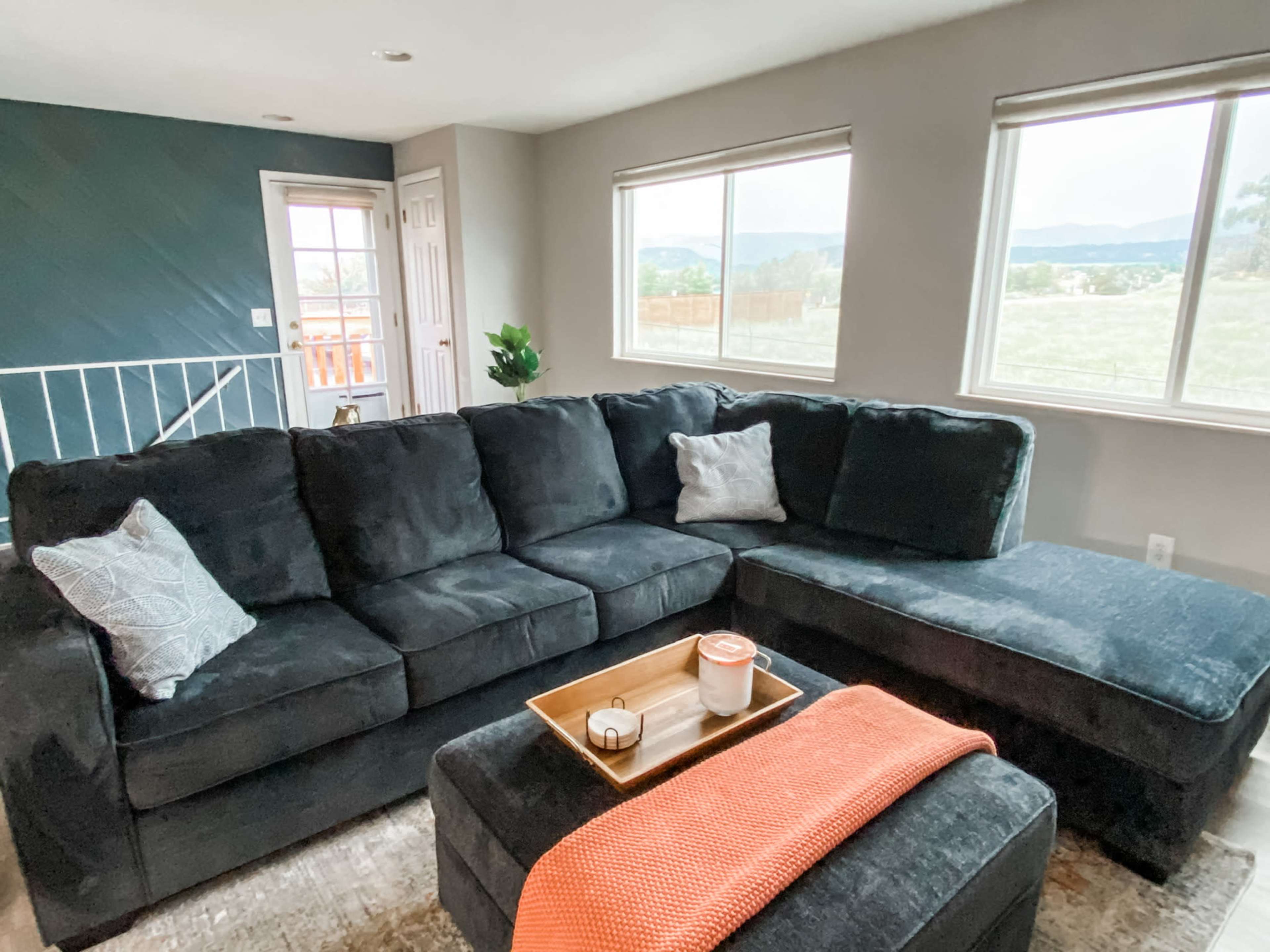 A dark sectional sofa with a throw blanket and tray on a coffee table, set in a bright living room with large windows and a view of the outdoors.
