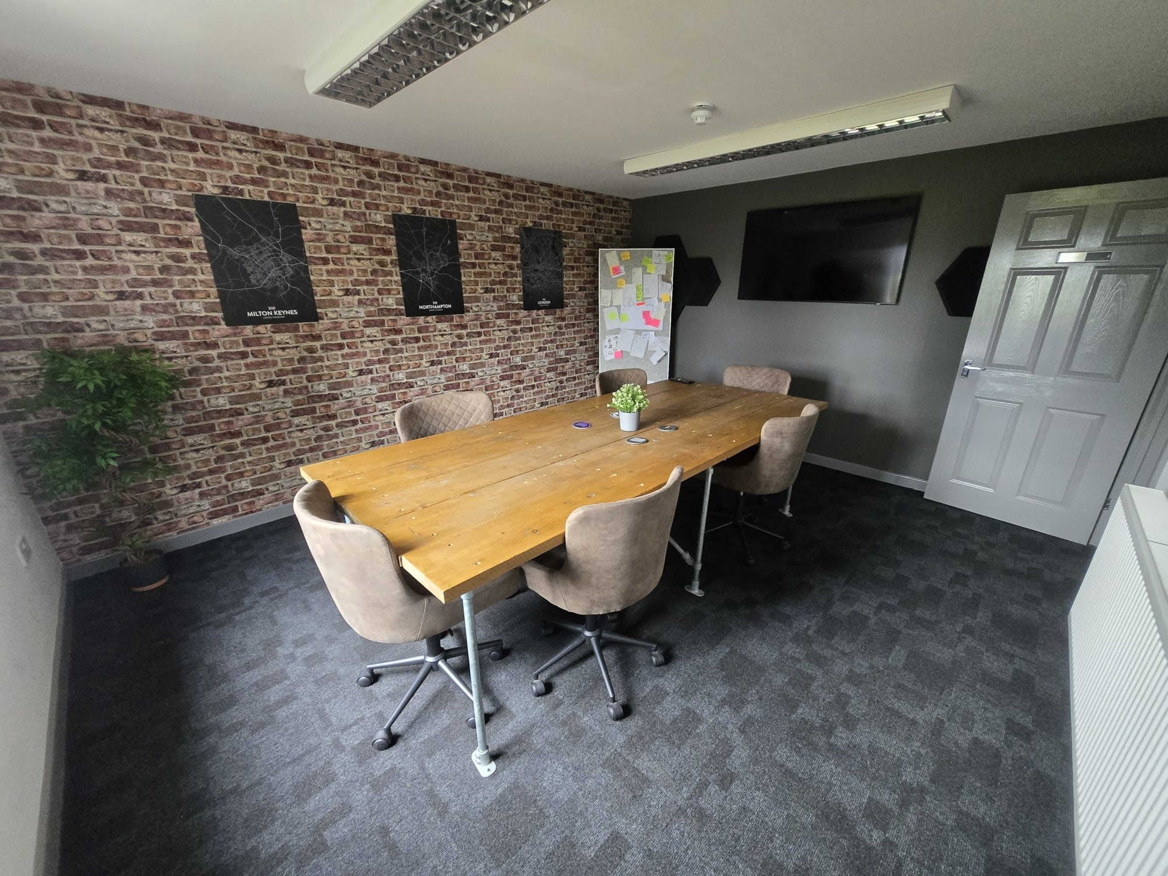 The image shows a meeting room with a wooden table surrounded by four chairs, a plant in one corner, and a television mounted on the wall, all against a backdrop of brick-patterned wallpaper and a grey wall.