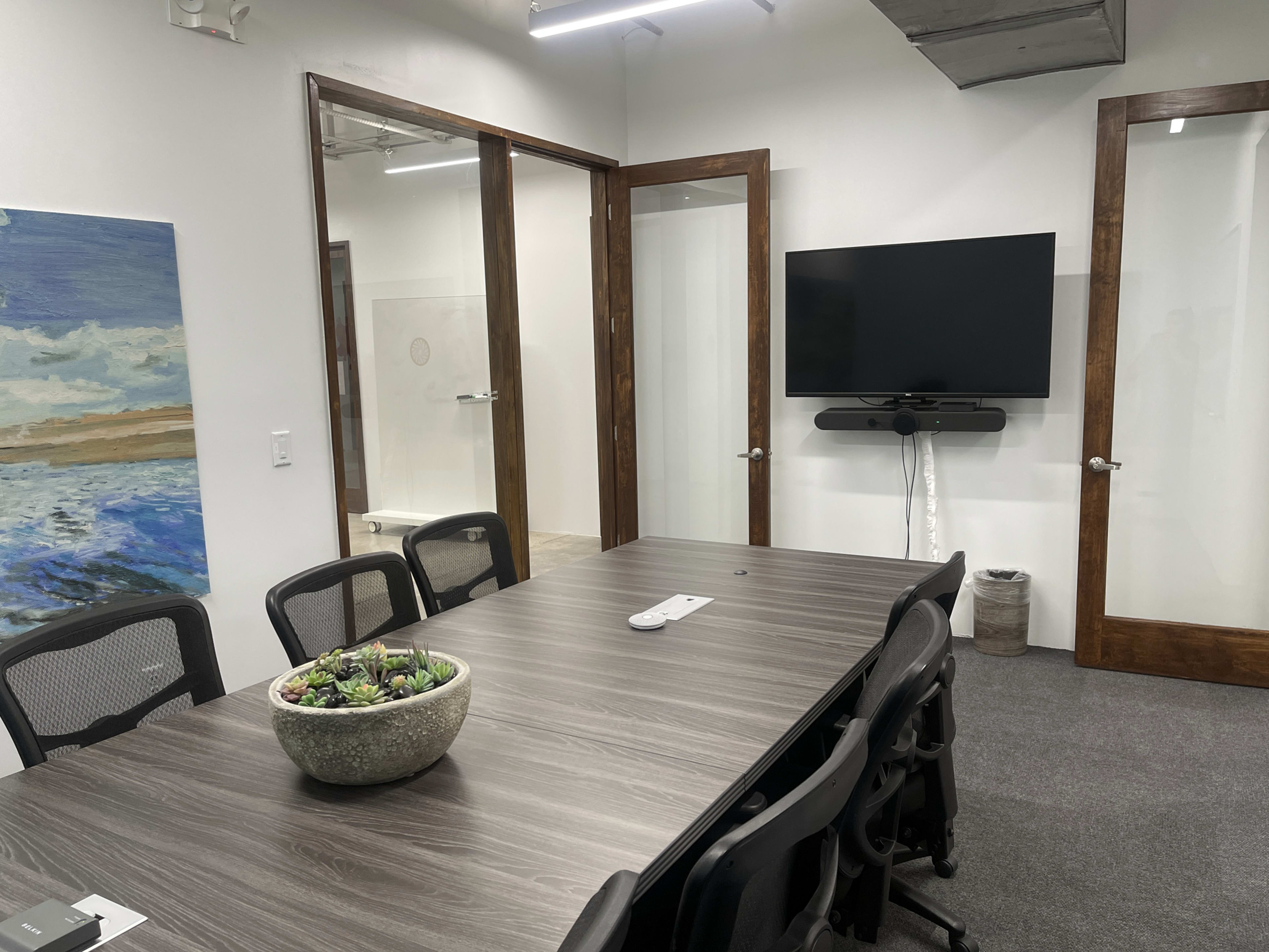 A conference room features a large wooden table, black chairs, a television mounted on the wall, and a potted plant in the center.