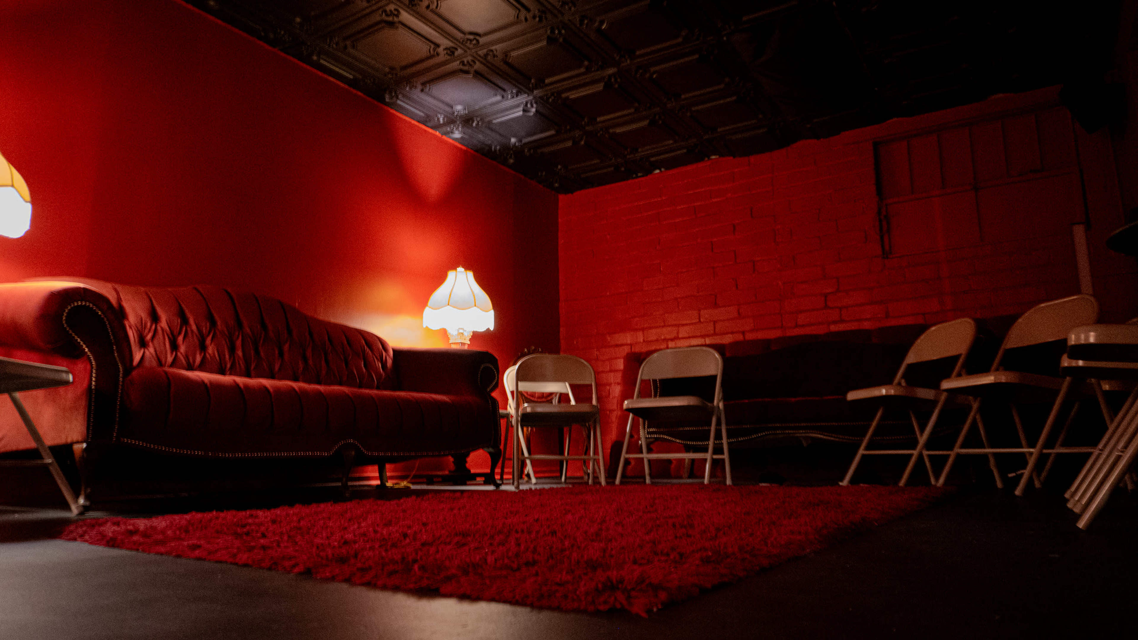 The image shows a dimly lit room with red walls, featuring a plush red couch, a lamp, and several white folding chairs arranged around a red rug.