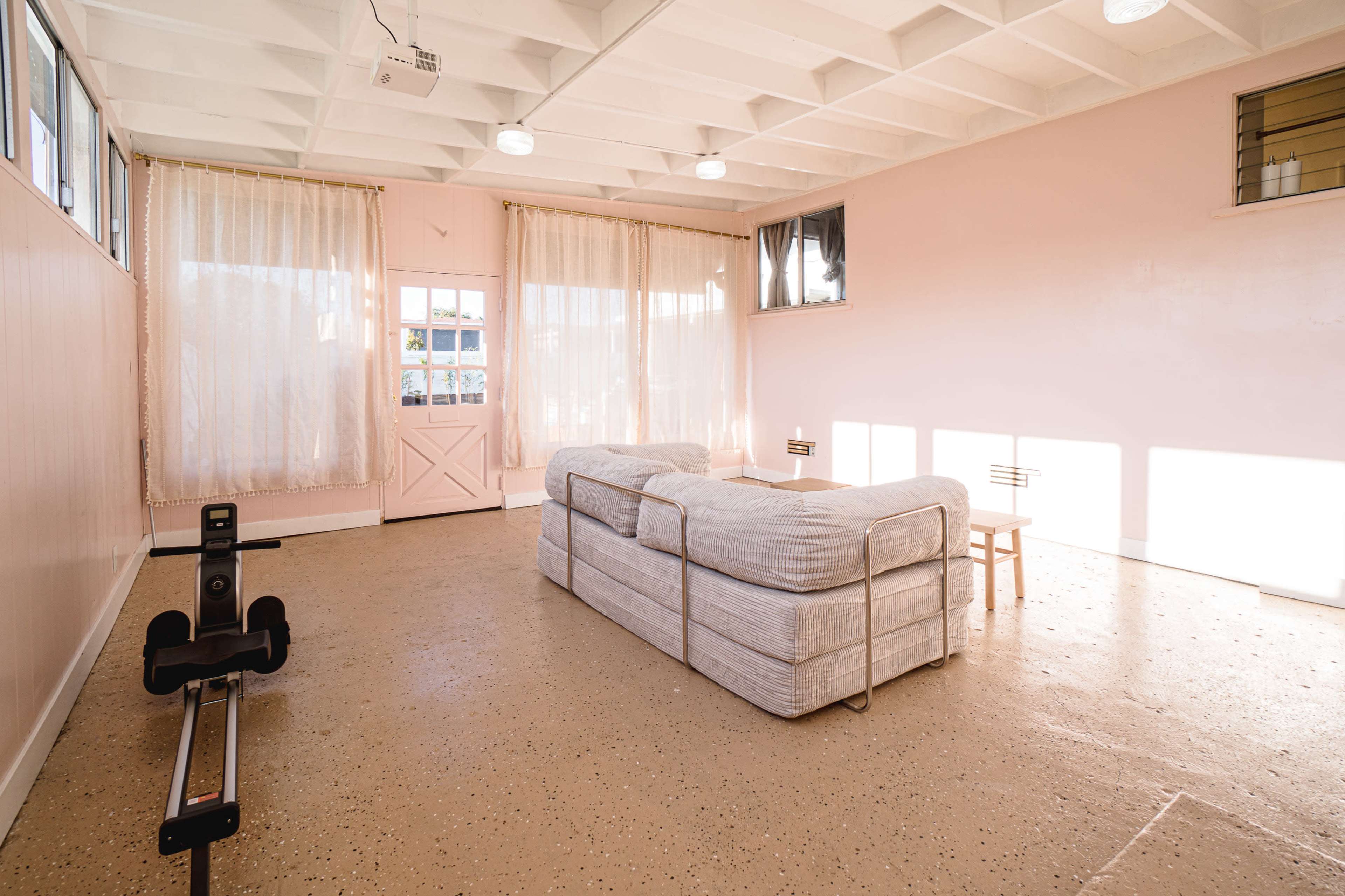 The image shows a minimalistic, light-filled room with a pink wall, a gray sofa, a small table, and two large windows draped with sheer curtains.