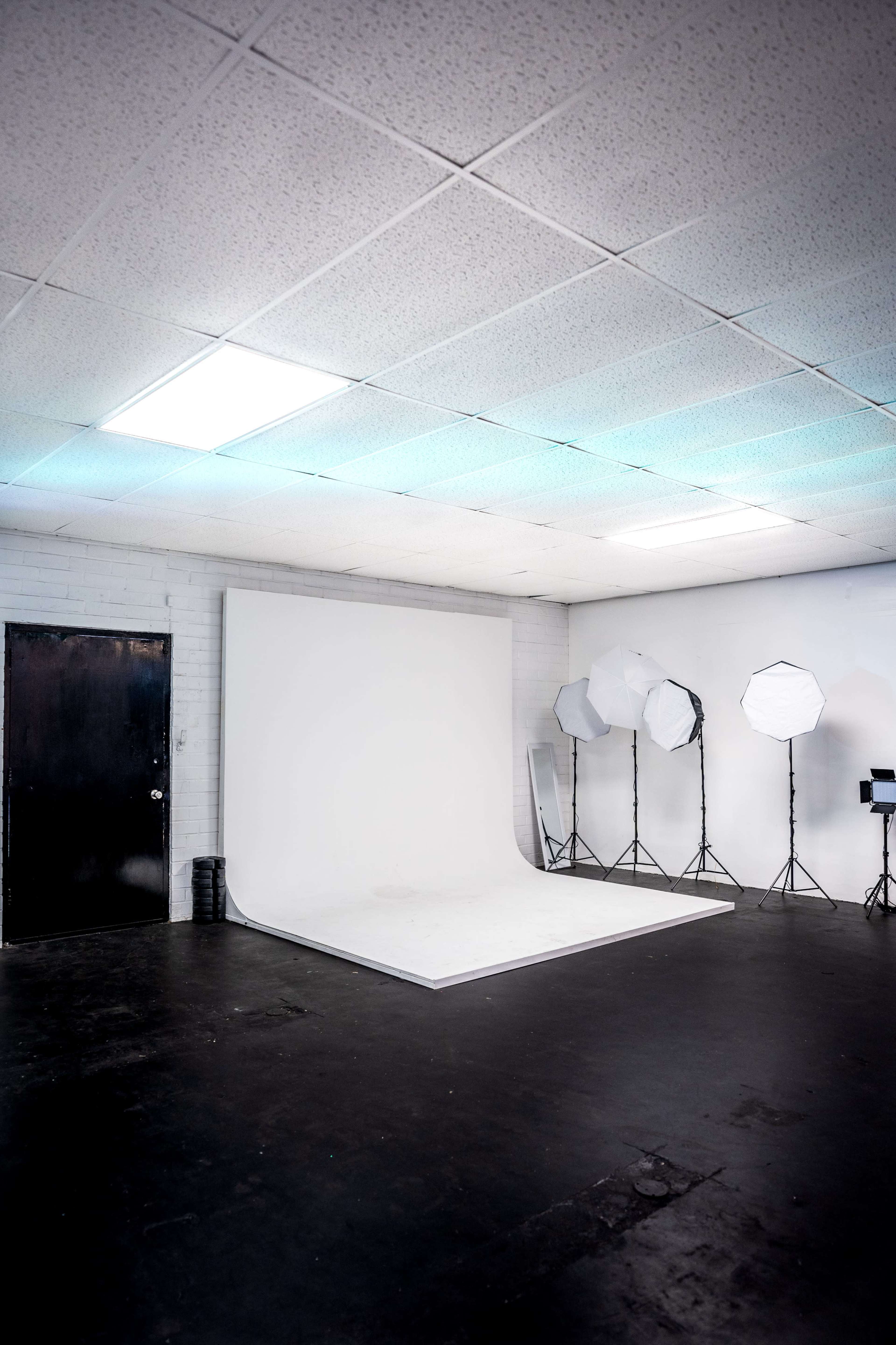 An empty photography studio with a white backdrop, black flooring, and lighting equipment set up on one side.