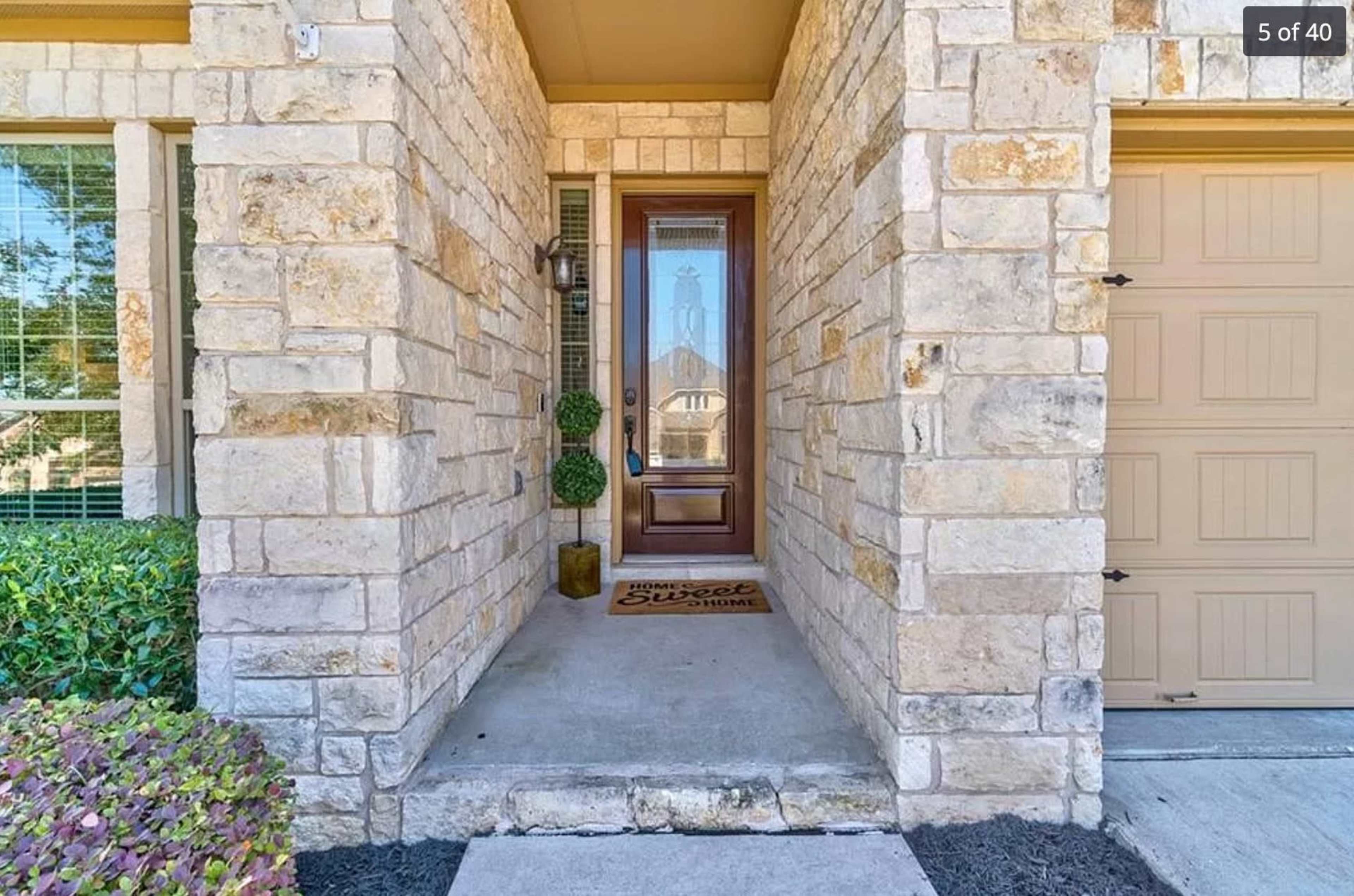 The entrance of a house featuring a stone facade, a wooden front door with a mat that reads "Home Sweet Home," and green potted plants on either side.
