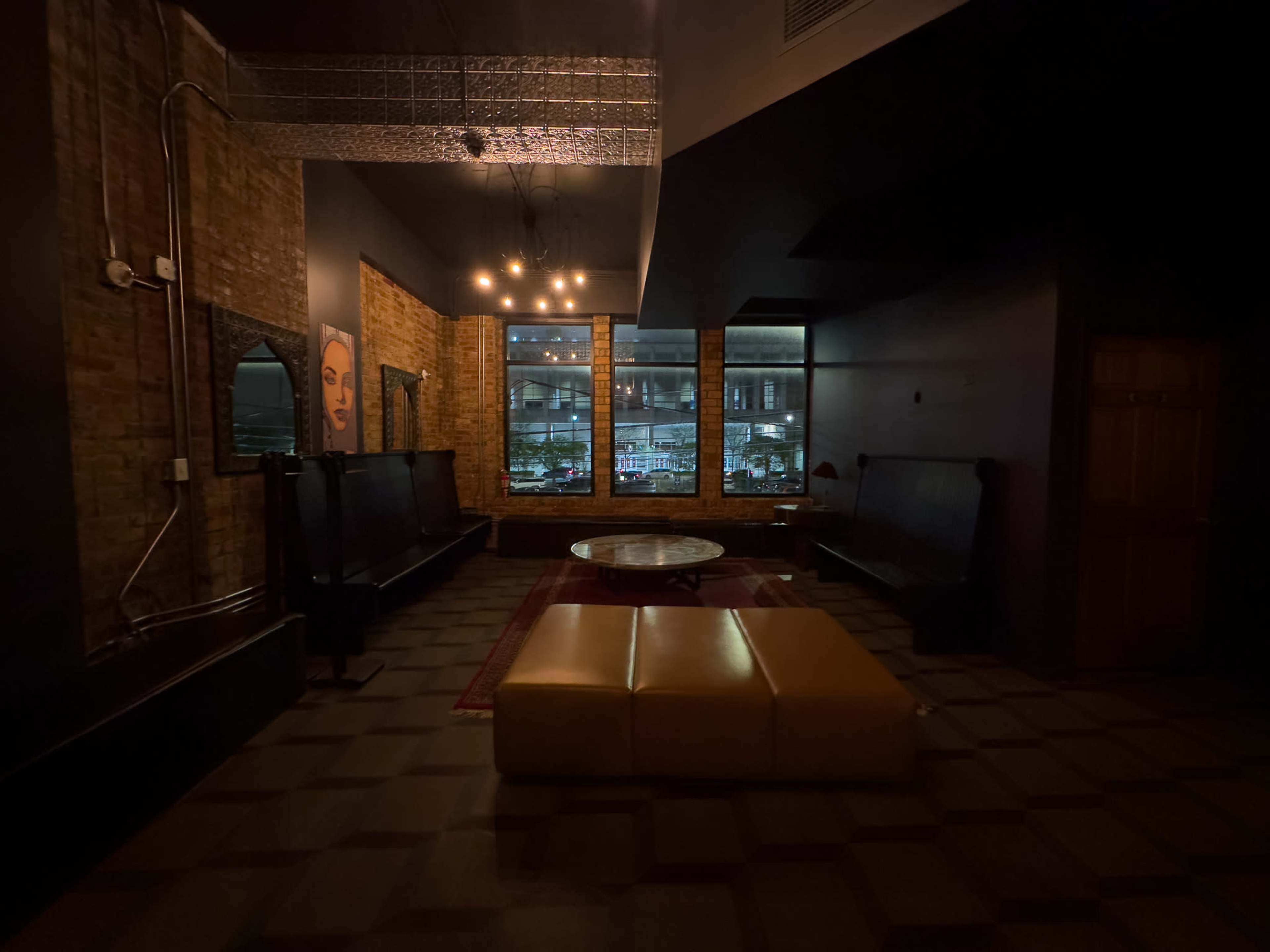 The image shows an empty lounge area with dark walls, a central coffee table, and large windows revealing a city view.