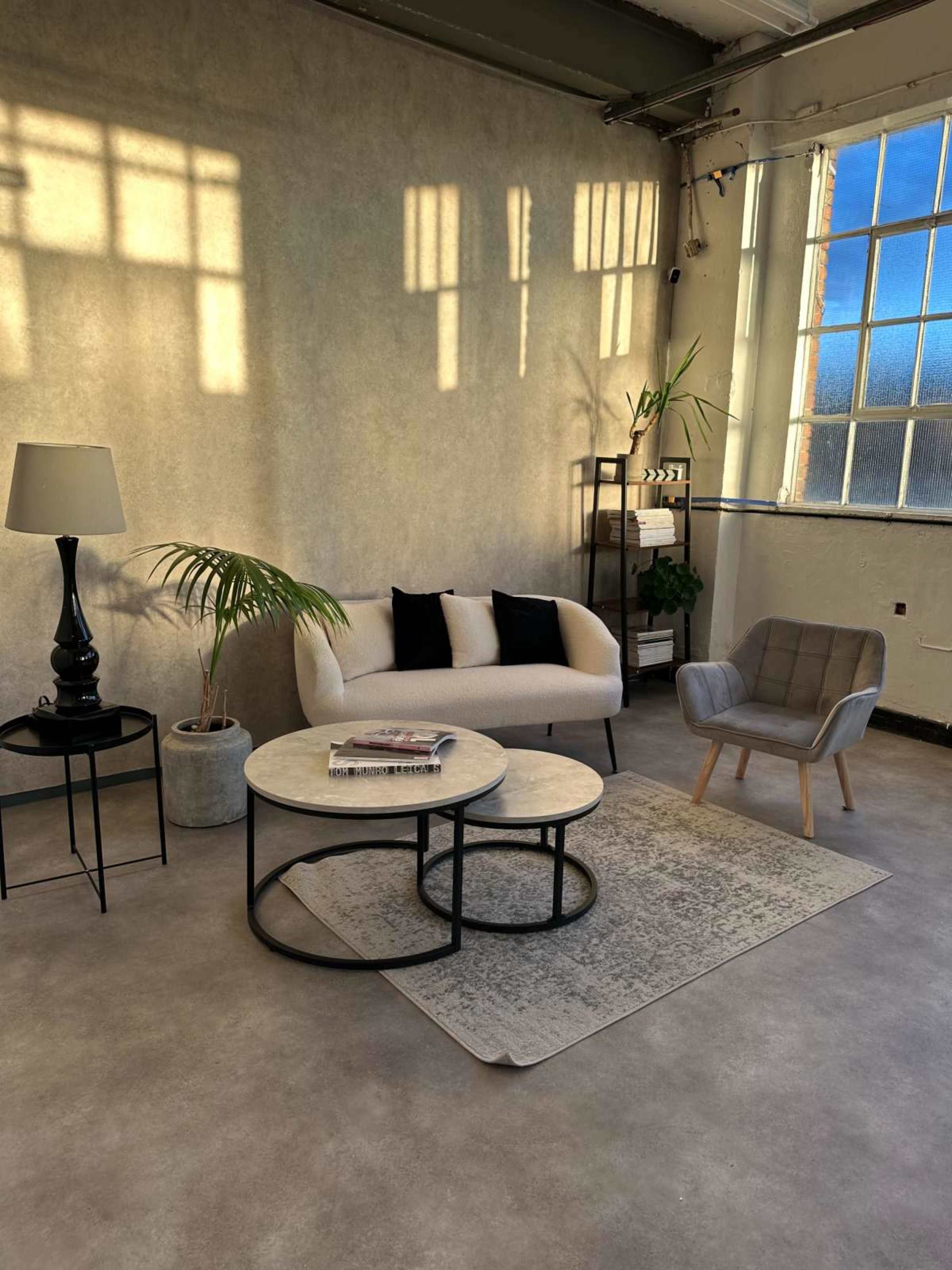 A living room scene features a light-colored sofa, two coffee tables, a lamp, a plant, and an armchair against a wall with large windows casting shadows.