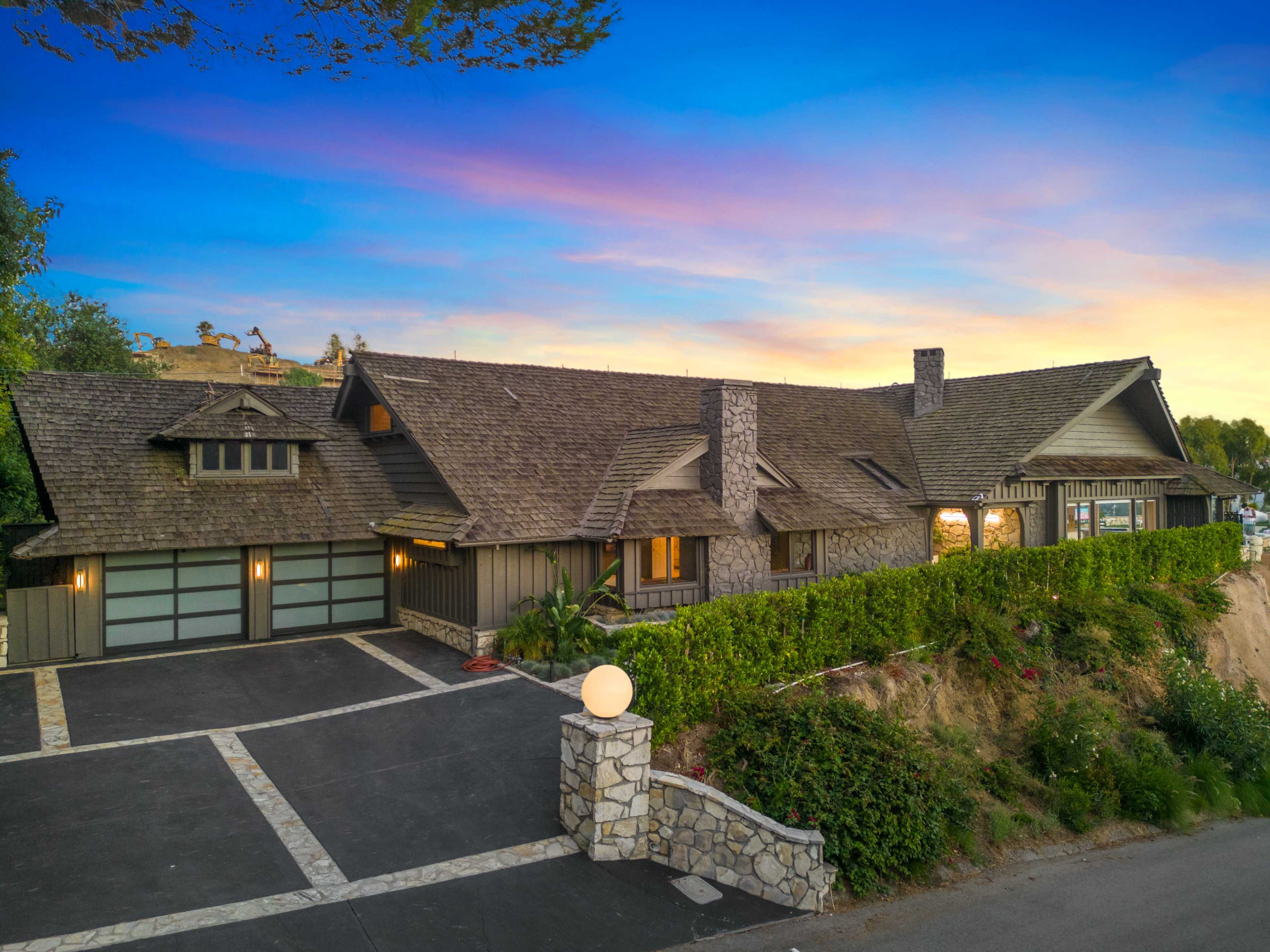 A large, single-story house with a stone and wood exterior, set against a sunset sky, with a paved driveway and landscaped front yard.