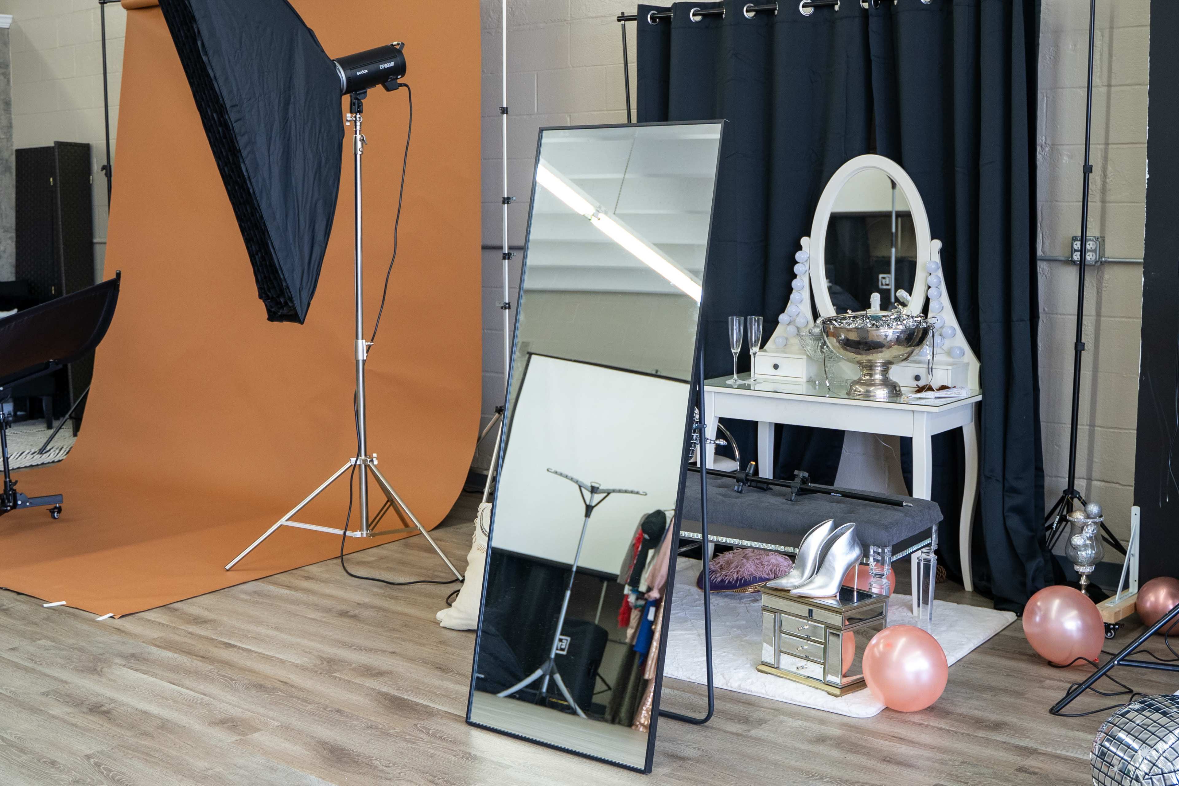 A photography studio setup with a large mirror, a lighting stand, a vintage-style dressing table, high heels, and decorative balloons on a wooden floor.