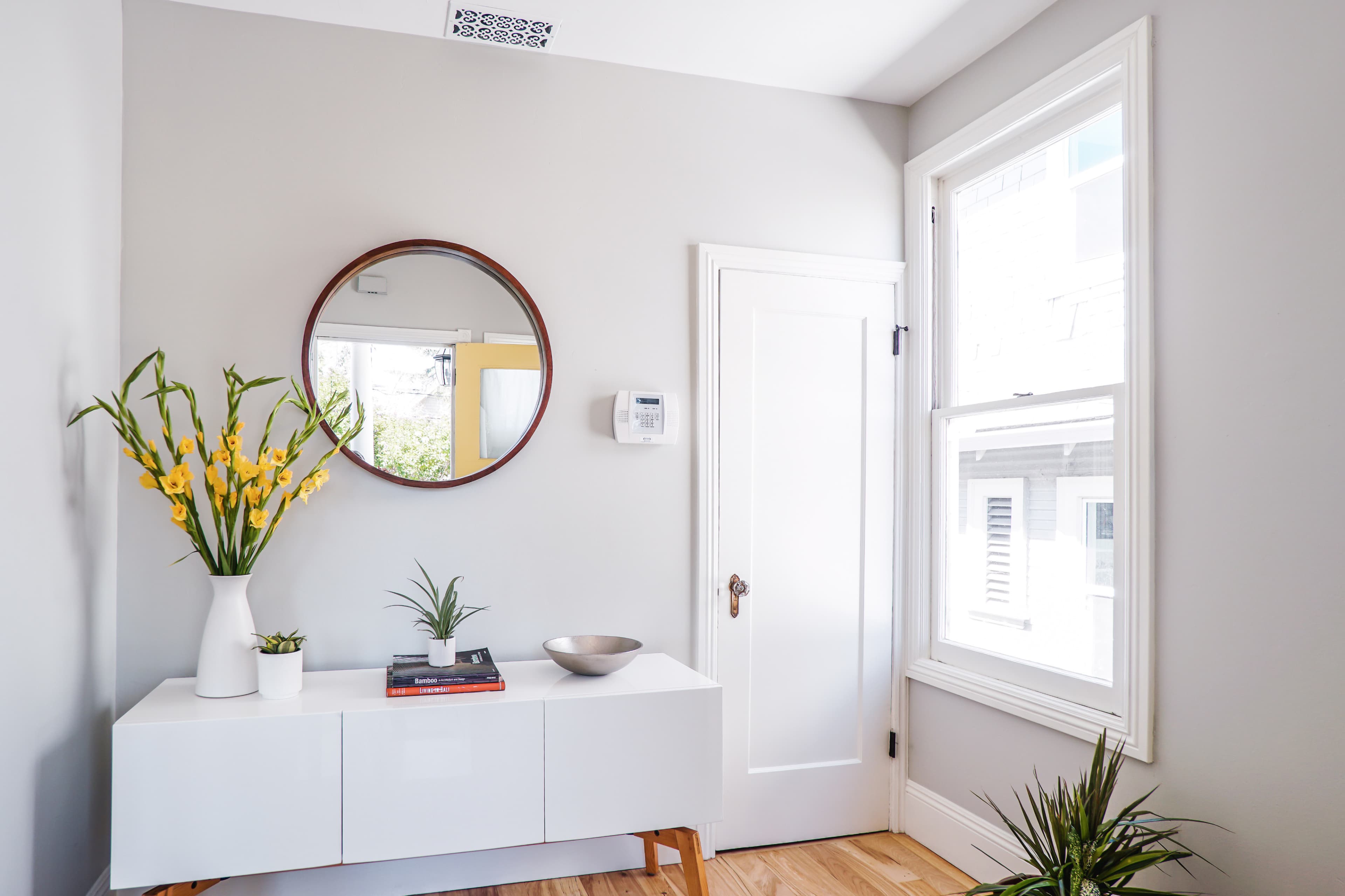 A bright entryway features a white console table, a round mirror, and potted plants beside a window.