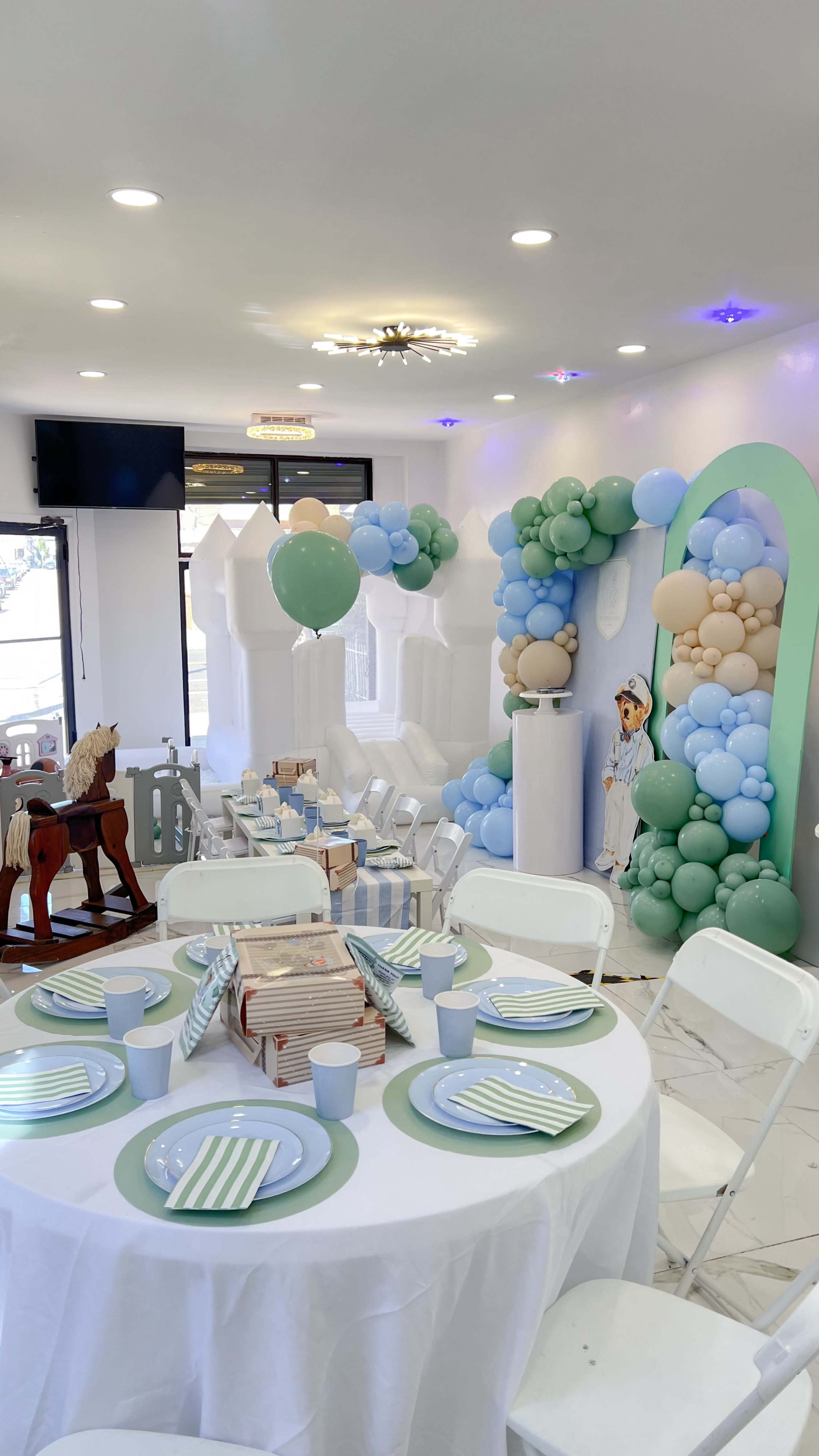 The image shows a decorated party space with a balloon arch, a backdrop featuring playful colors, and tables set with plates and cups.
