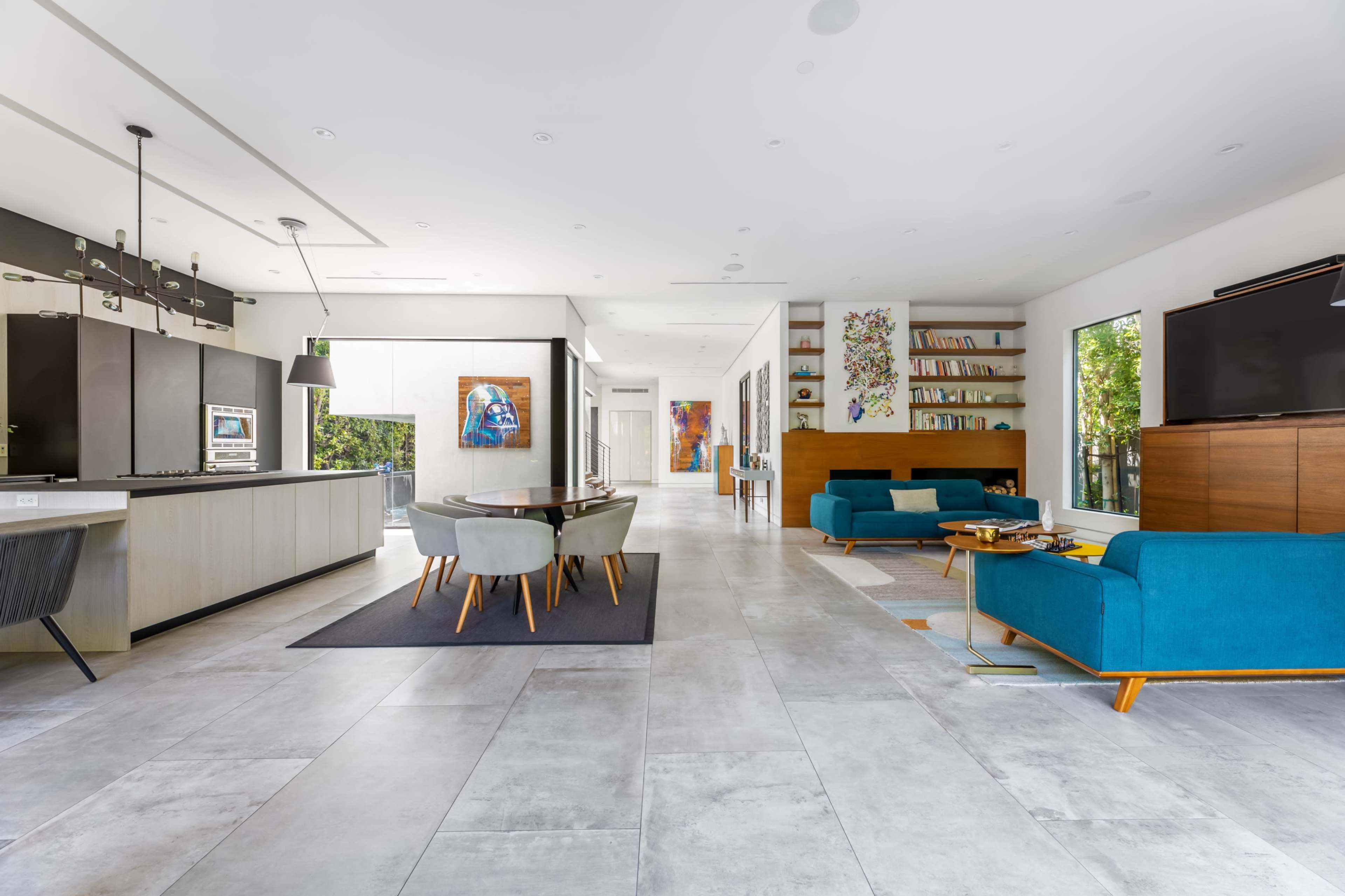 The image shows a modern, open-concept living area featuring a dining table, teal sofas, and a variety of colorful artwork on the walls.