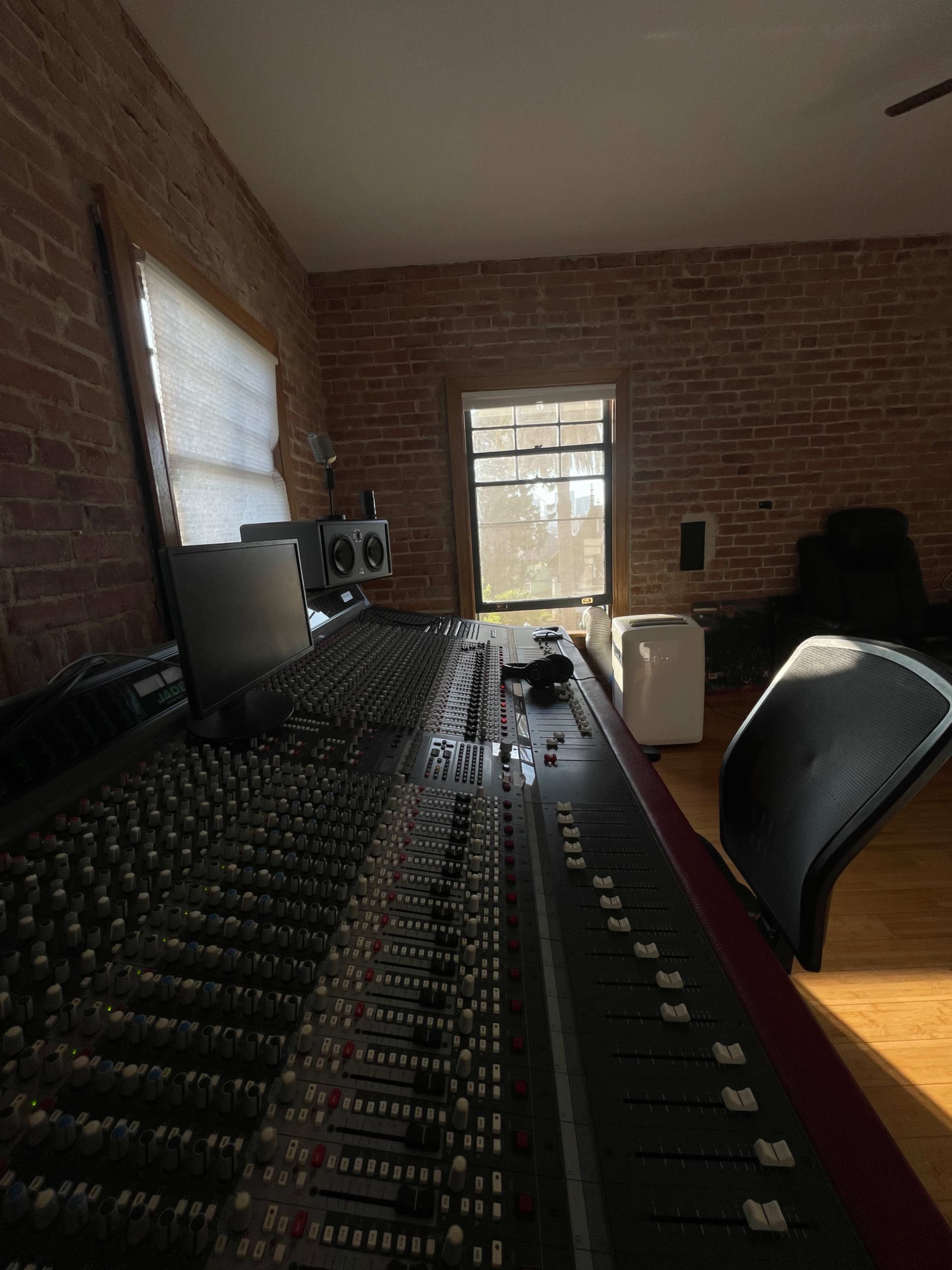 A recording studio with a large mixing console, computer monitor, and a chair, overlooking a window with a view outside.
