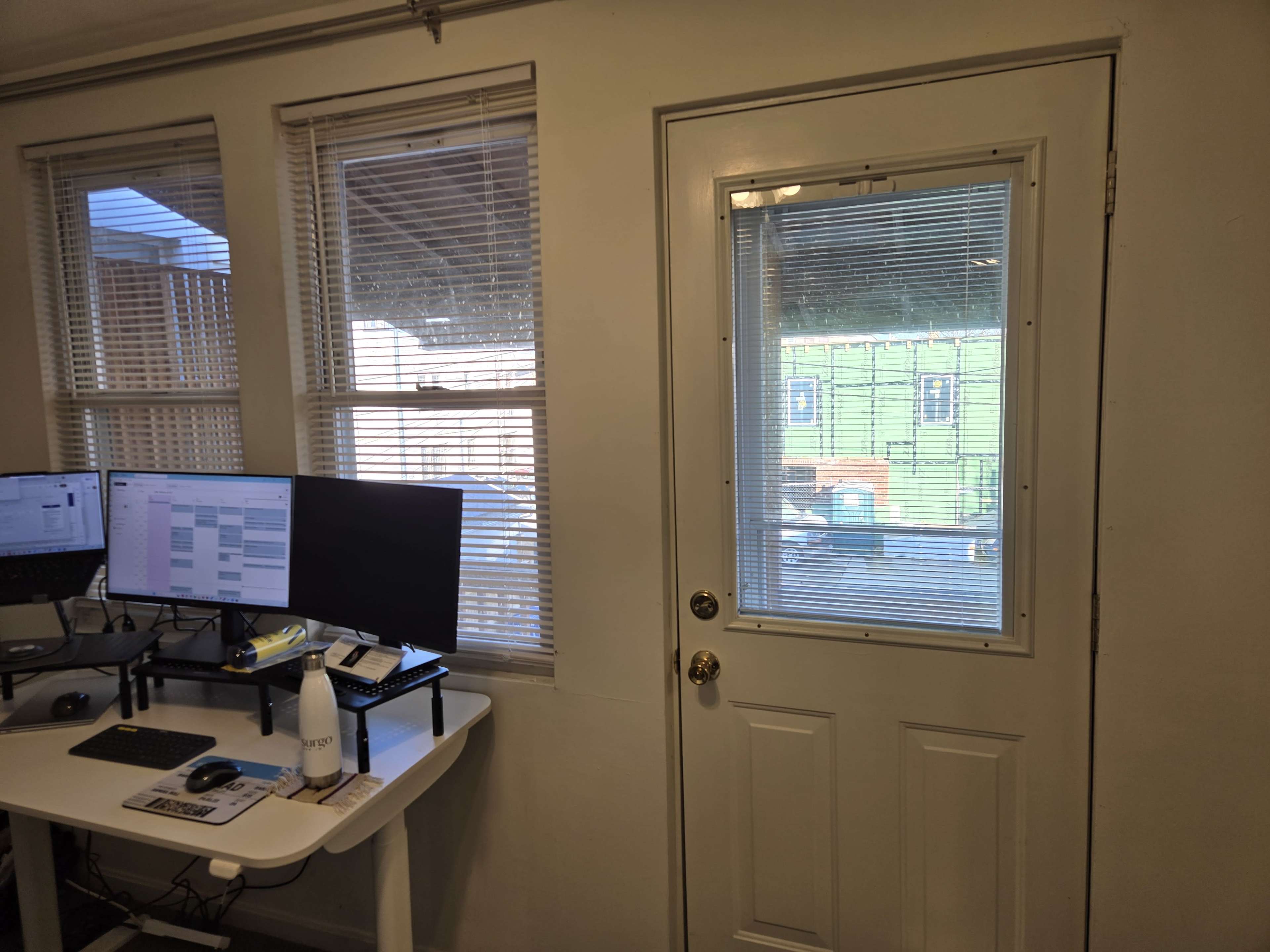 The image shows an interior view of a room with a door that has a window, two computer monitors on a desk, and blinds on the windows.