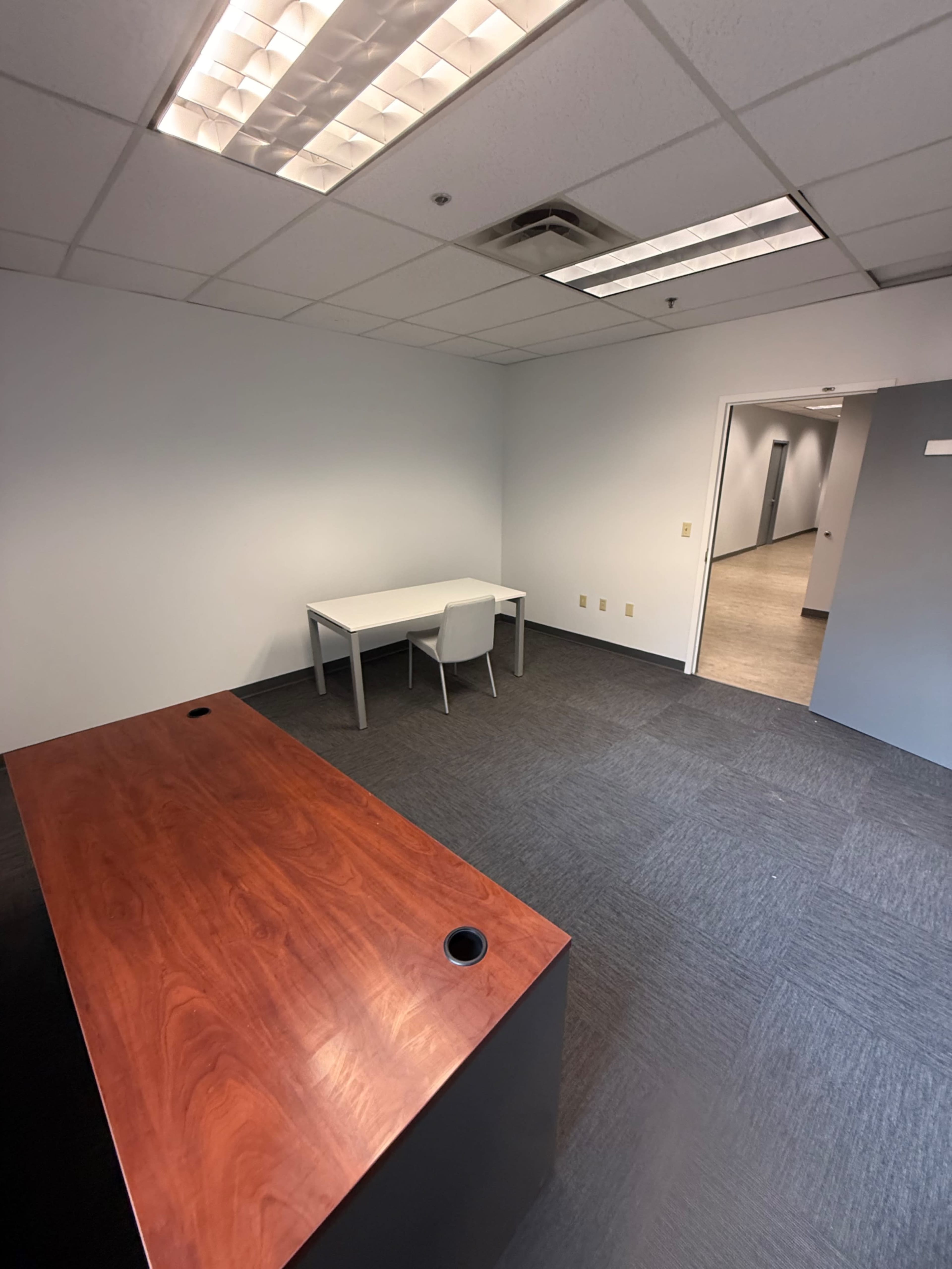 An office space features a wooden desk and a white chair, with gray carpeting and a doorway leading to a hallway.
