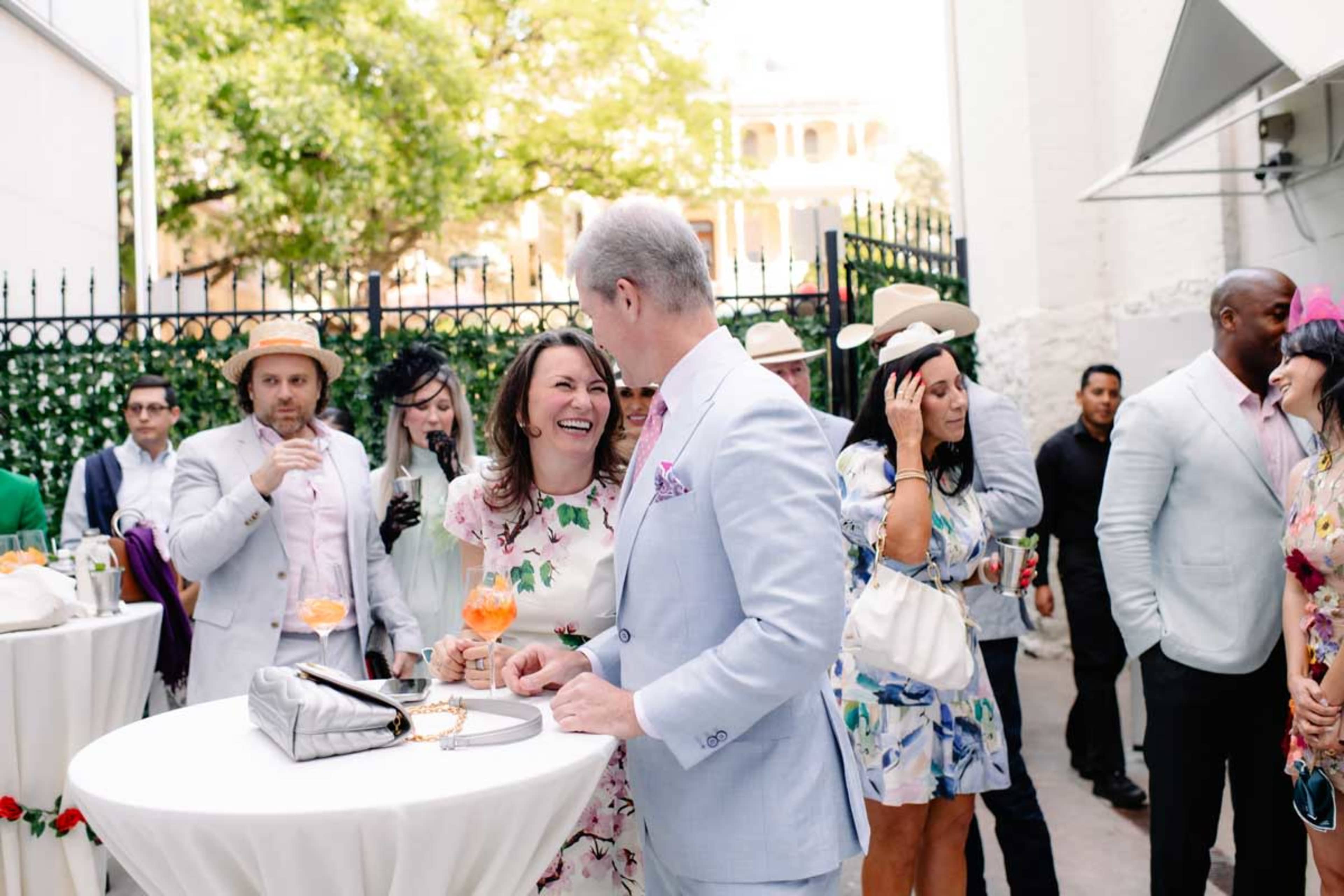 A group of elegantly dressed people socialize at a festive outdoor event with tables, drinks, and a backdrop of greenery and architectural details.