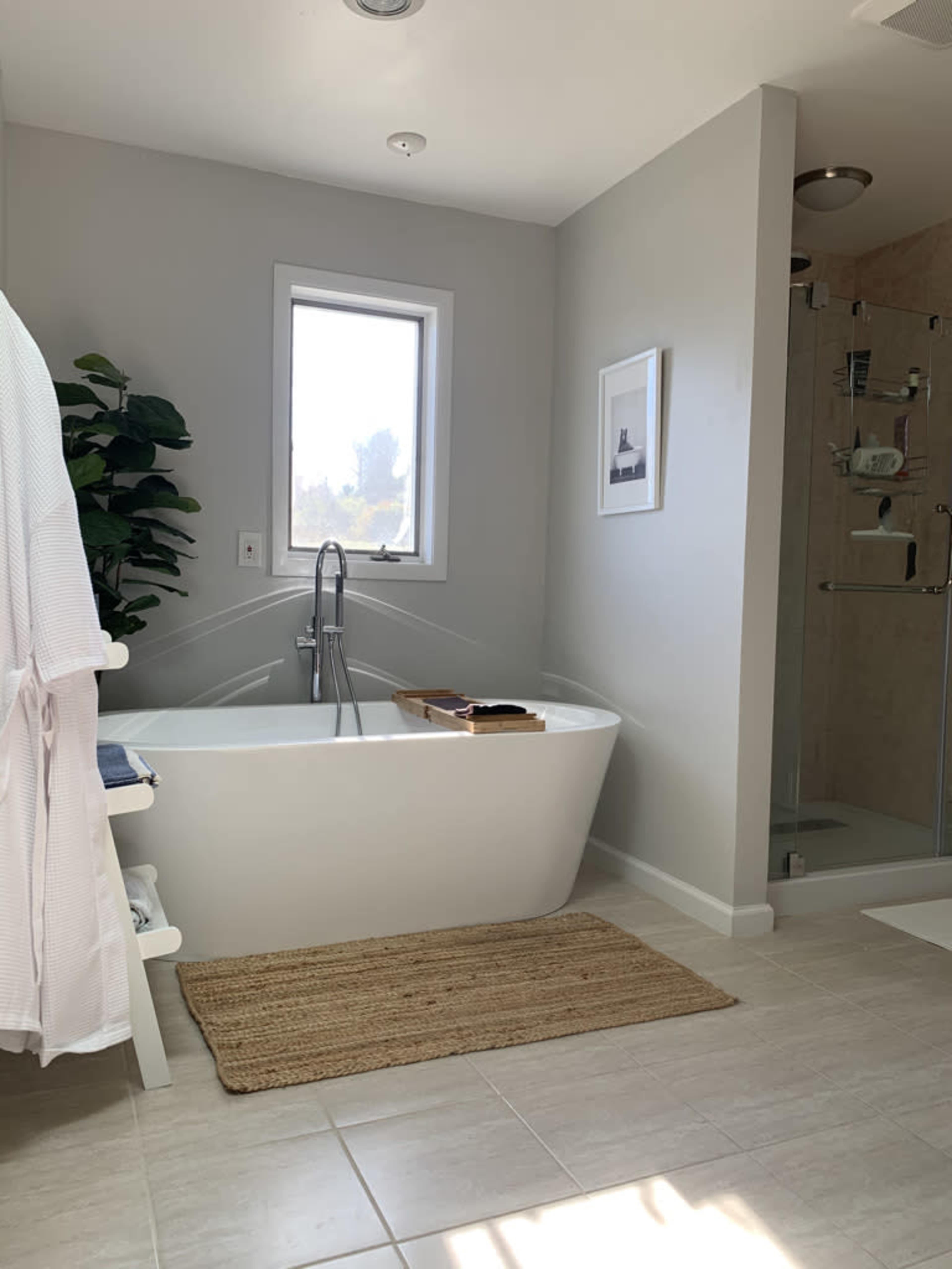 A modern bathroom featuring a freestanding bathtub, a glass shower enclosure, a window, and a woven rug on tiled flooring.