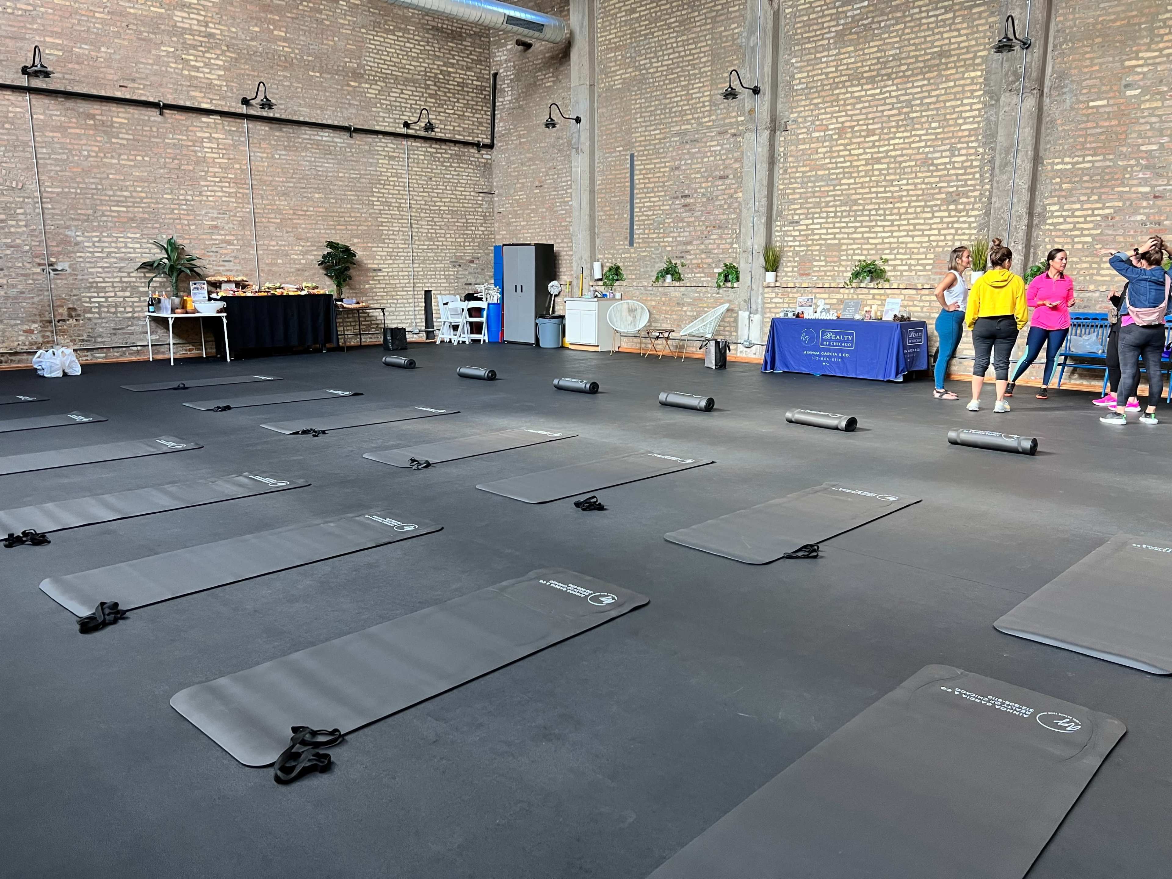 A spacious room with exercise mats arranged on a black floor, while a group of people gathers near a table with refreshments in the background.