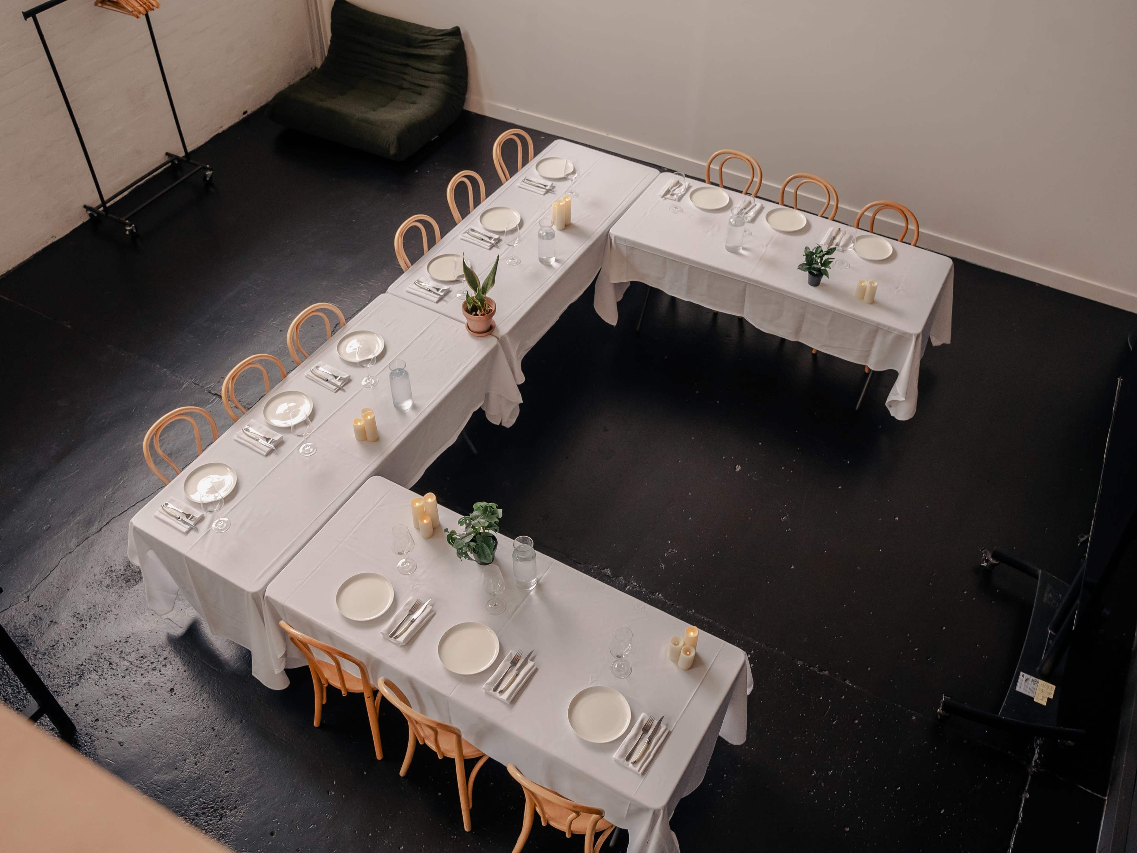 The image shows a dining setup with multiple tables arranged in a U-shape, covered with white tablecloths and set with plates, cutlery, and small plants.