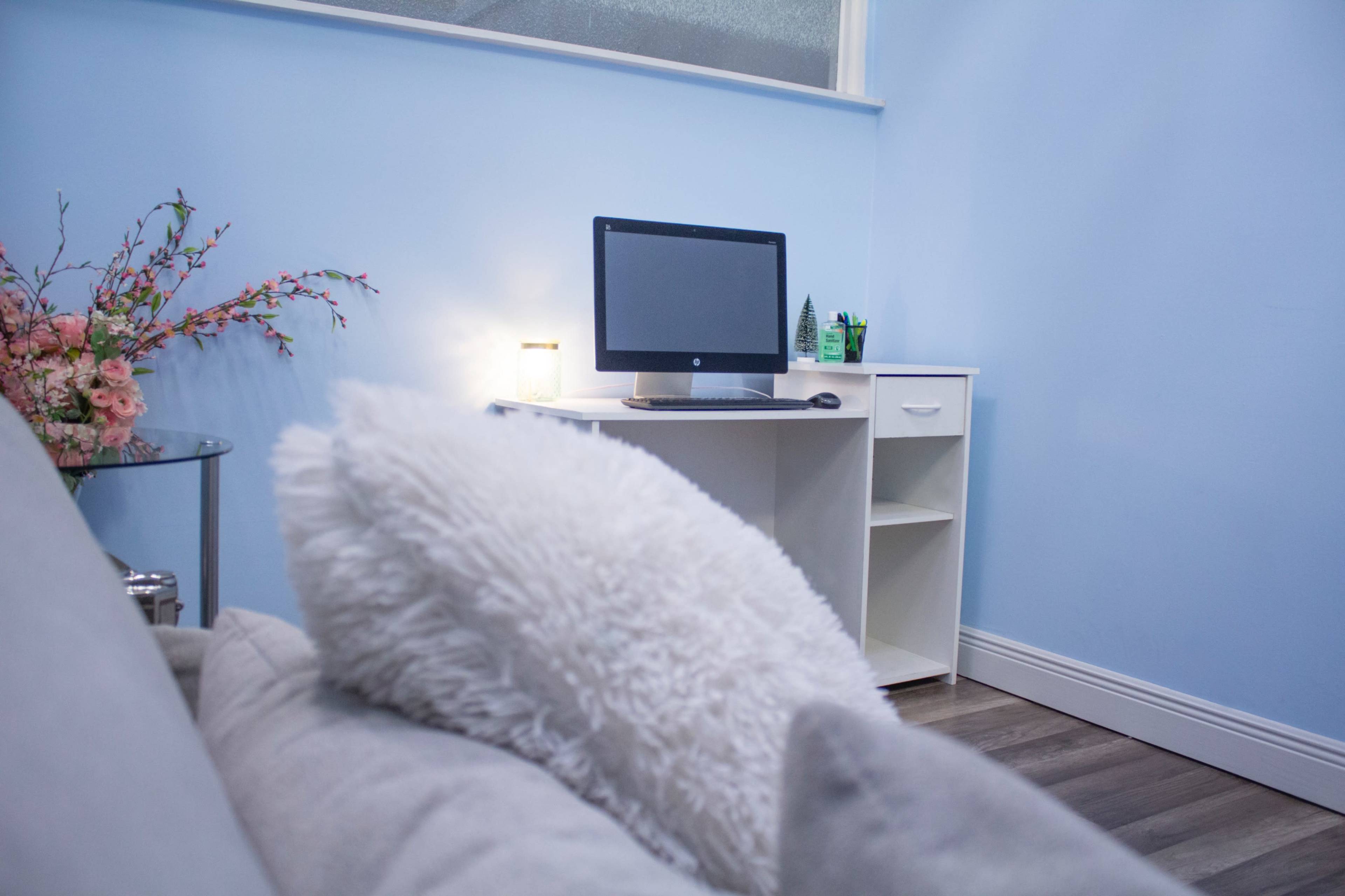 A light blue room with a computer on a white desk, a decorative plant, and a floral arrangement next to a gray sofa.