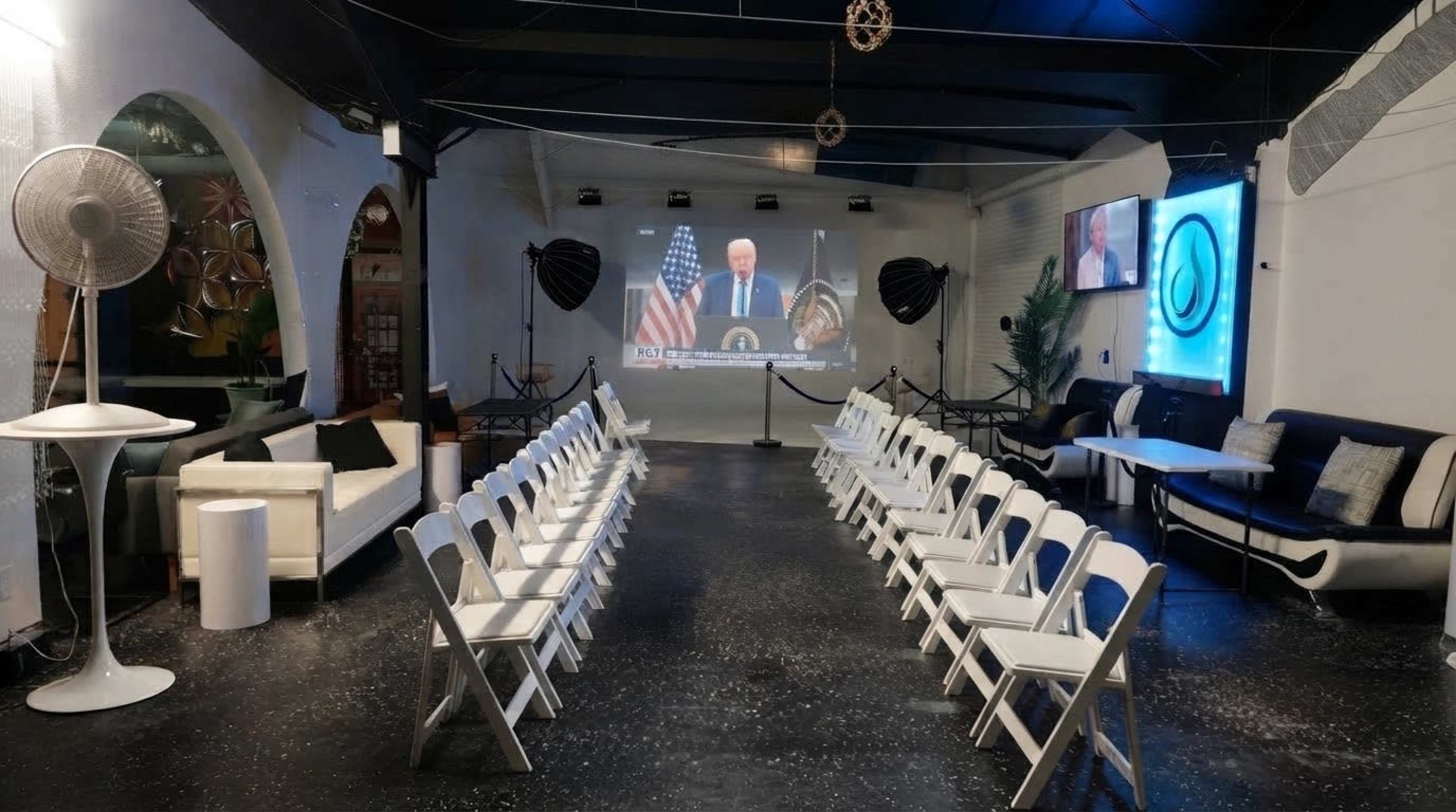 The image shows a minimalist indoor space set up for an event, featuring rows of white chairs facing a large screen displaying a speaker, with additional seating and decorative elements in the background.