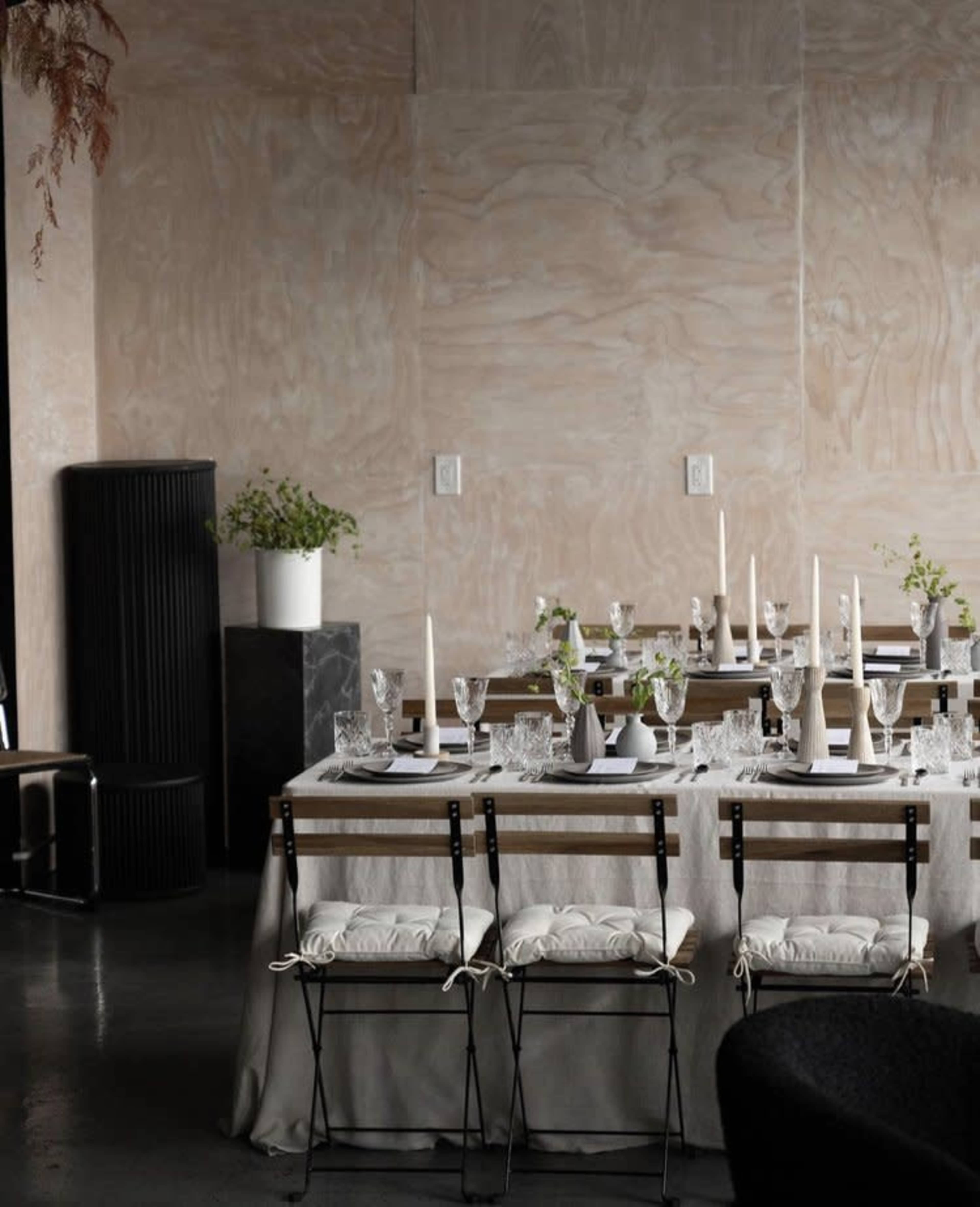 A dining table set with white tableware and glassware is arranged in a minimalist room with a wooden wall and greenery.