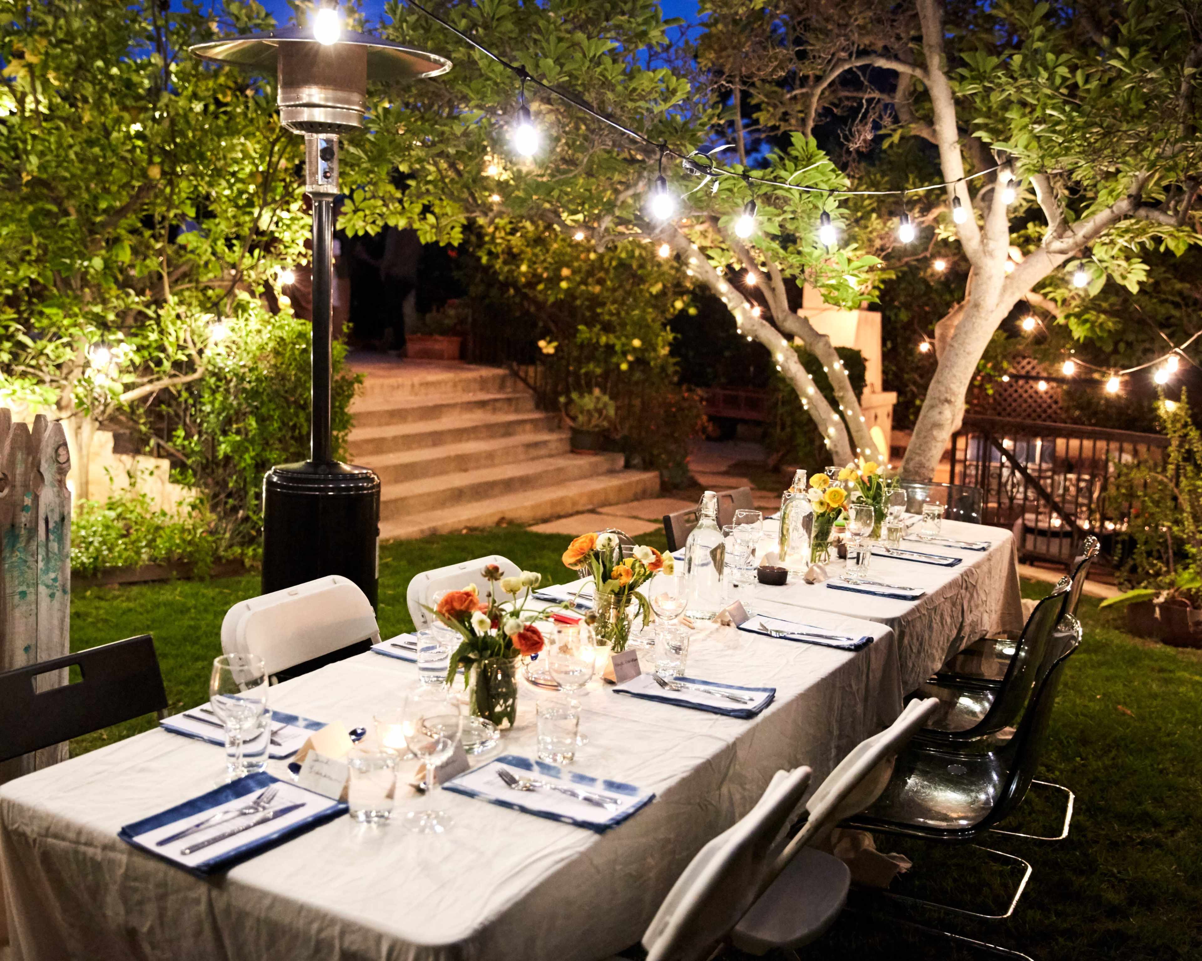 A long dining table is set outdoors, illuminated by string lights and surrounded by greenery.