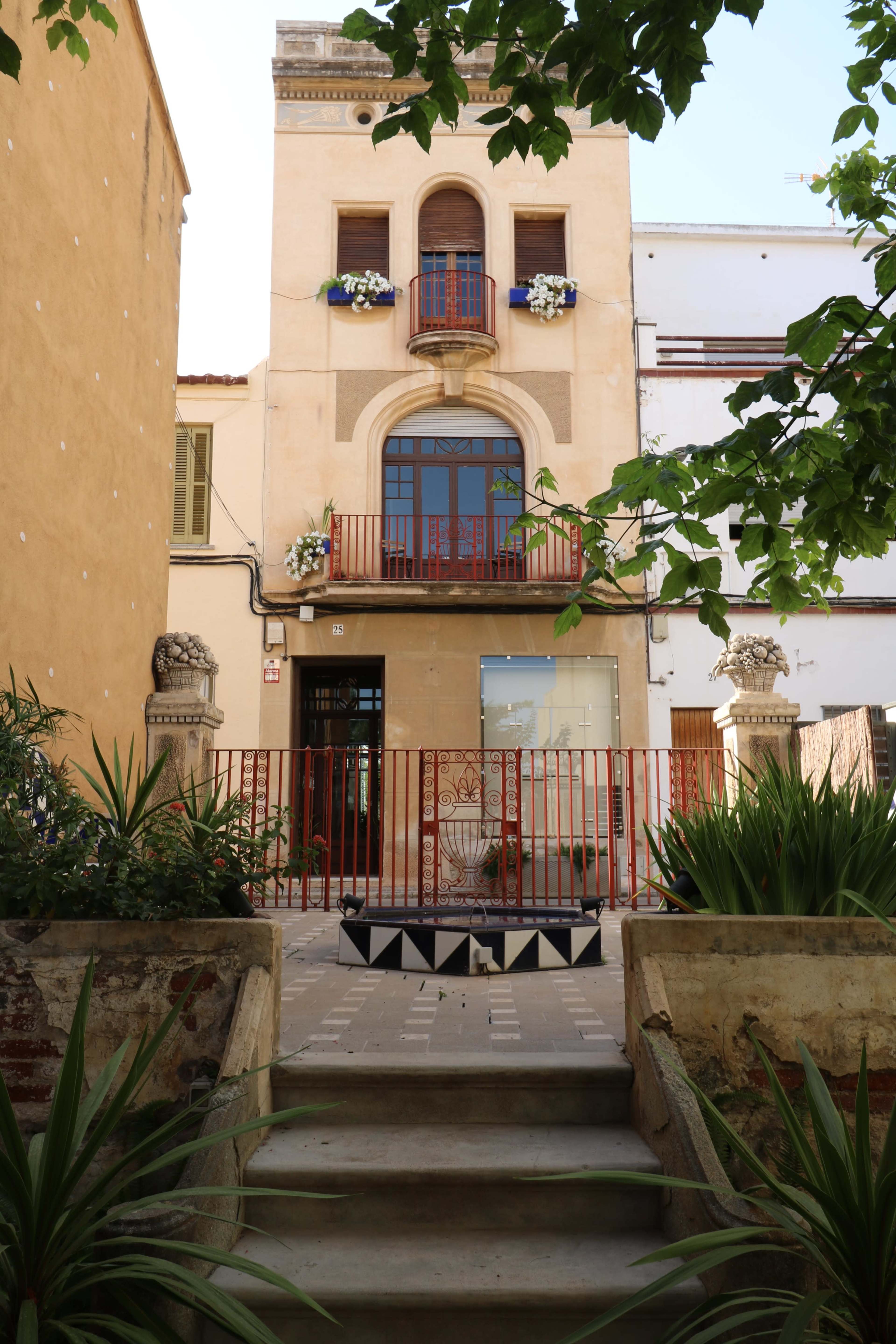 A three-story building with ornate architecture, a red balcony railing, and decorative plants in a courtyard setting.