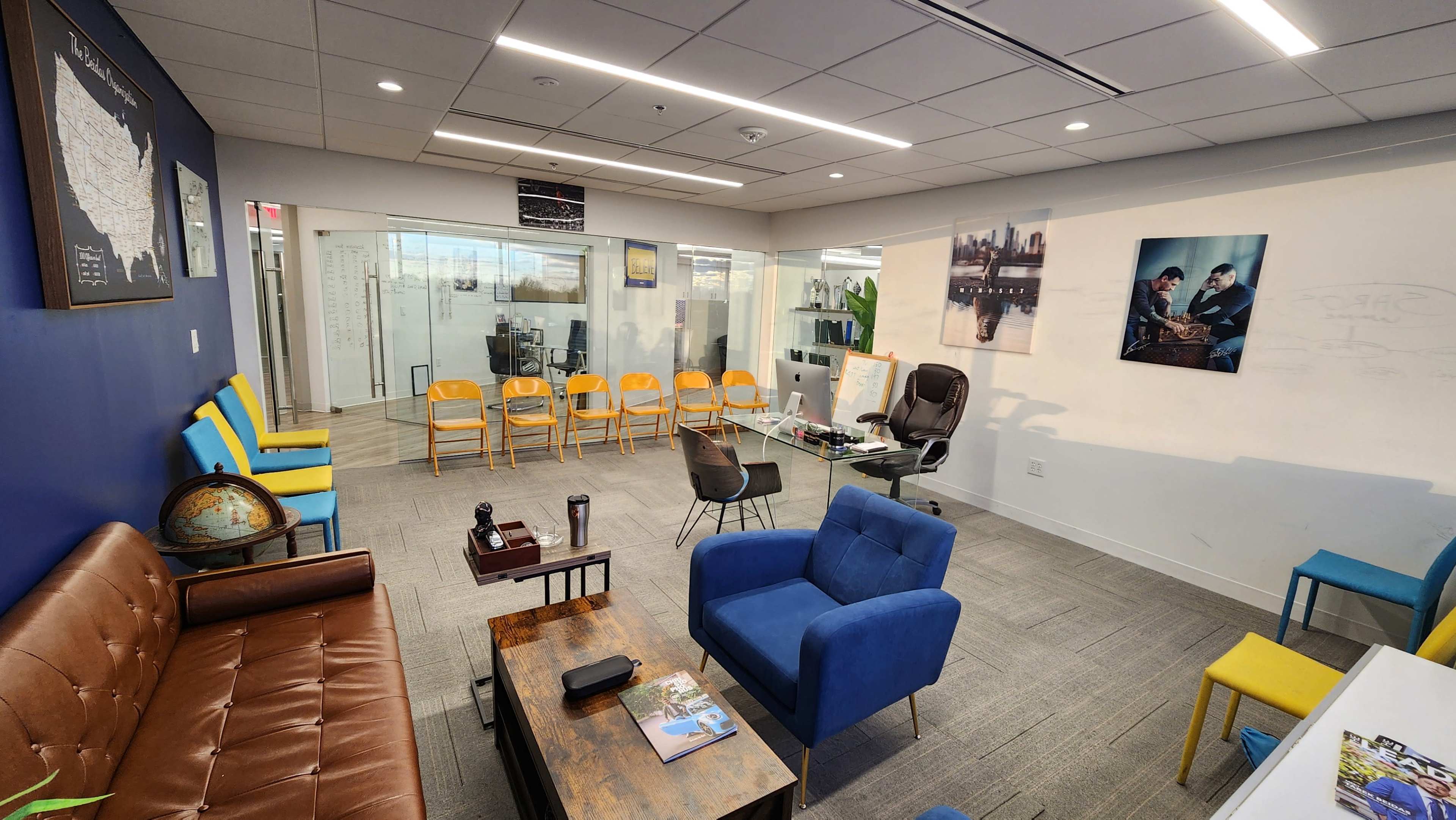 A modern office space with seating areas, a glass-walled meeting room, and colorful chairs arranged in rows.