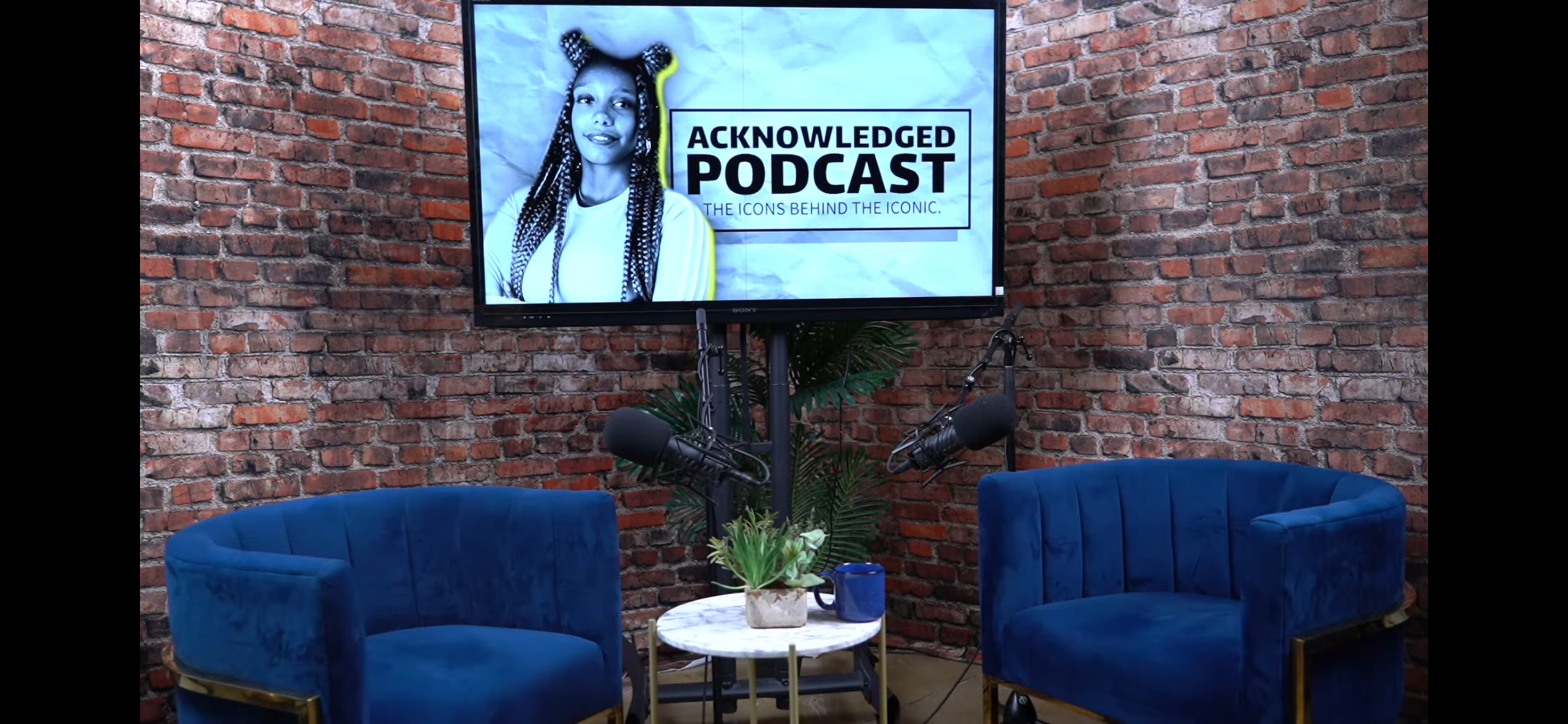 The image shows a podcast studio with two blue chairs, a small round table with a plant, and a wall-mounted screen displaying the title "ACKNOWLEDGED PODCAST" alongside a woman's portrait.