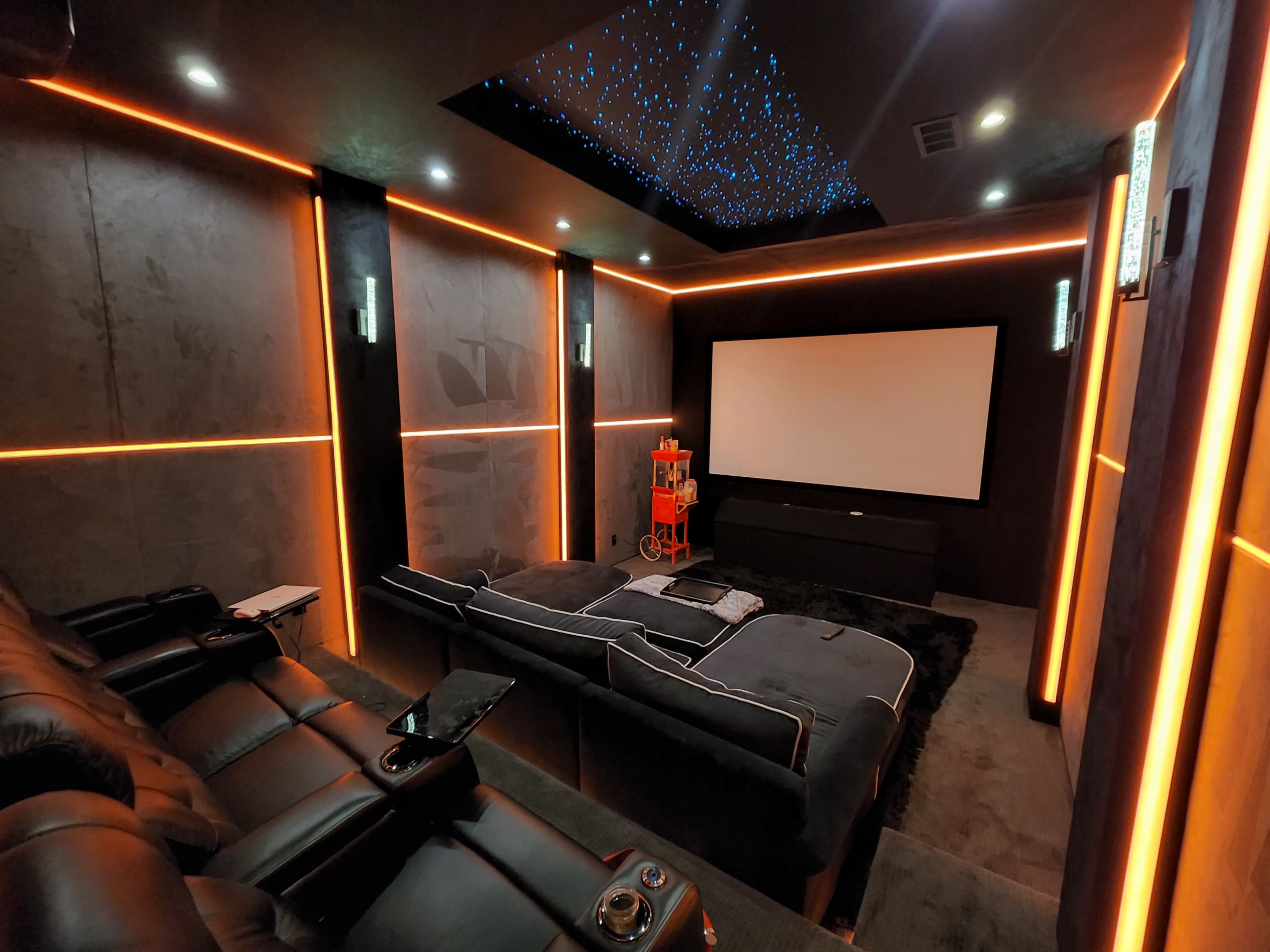 The image shows a modern home theater with black seating, orange accent lighting, and a large white screen.