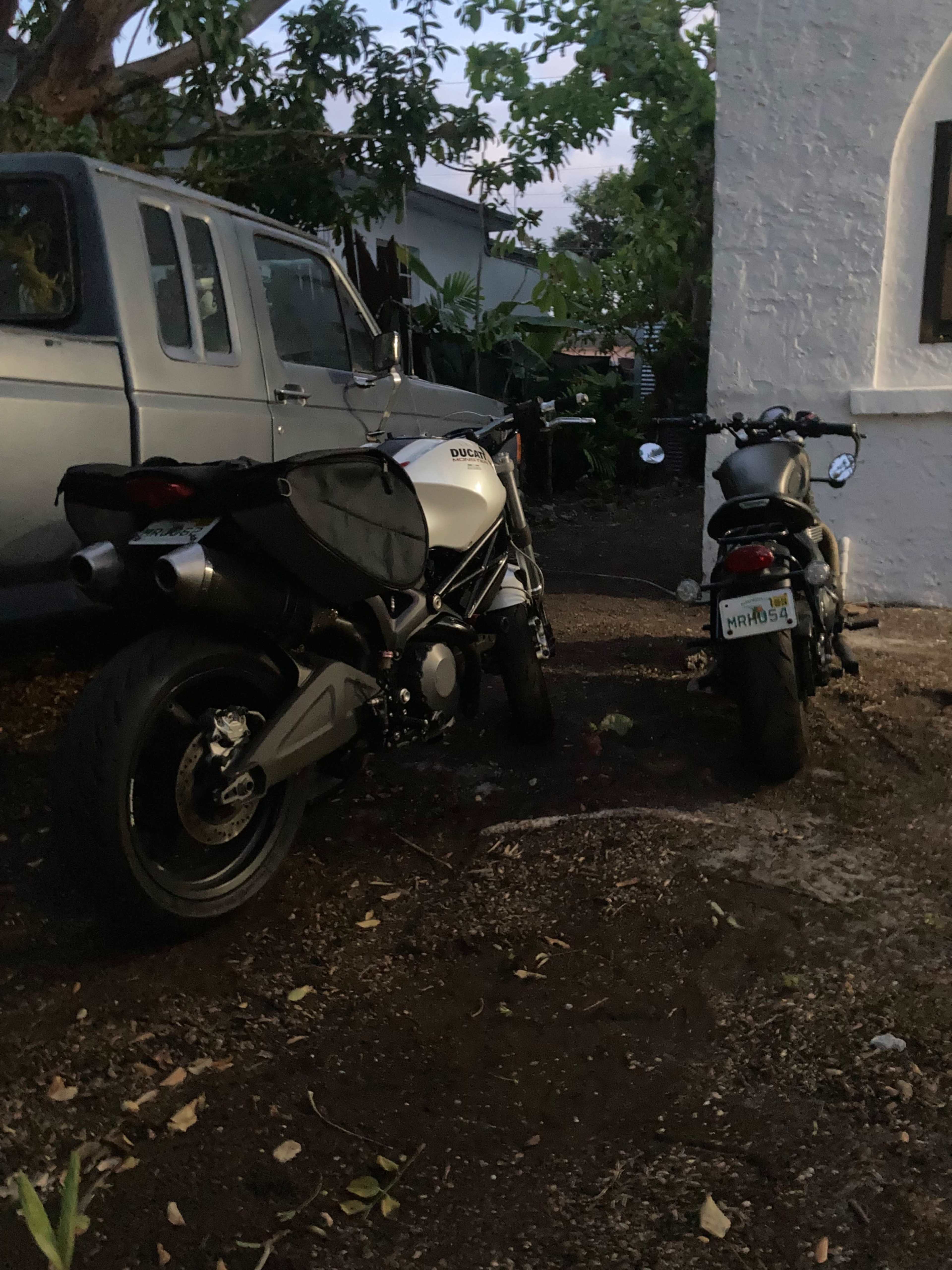 Two motorcycles are parked side by side in a narrow space beside a house, with a gray SUV visible in the background.