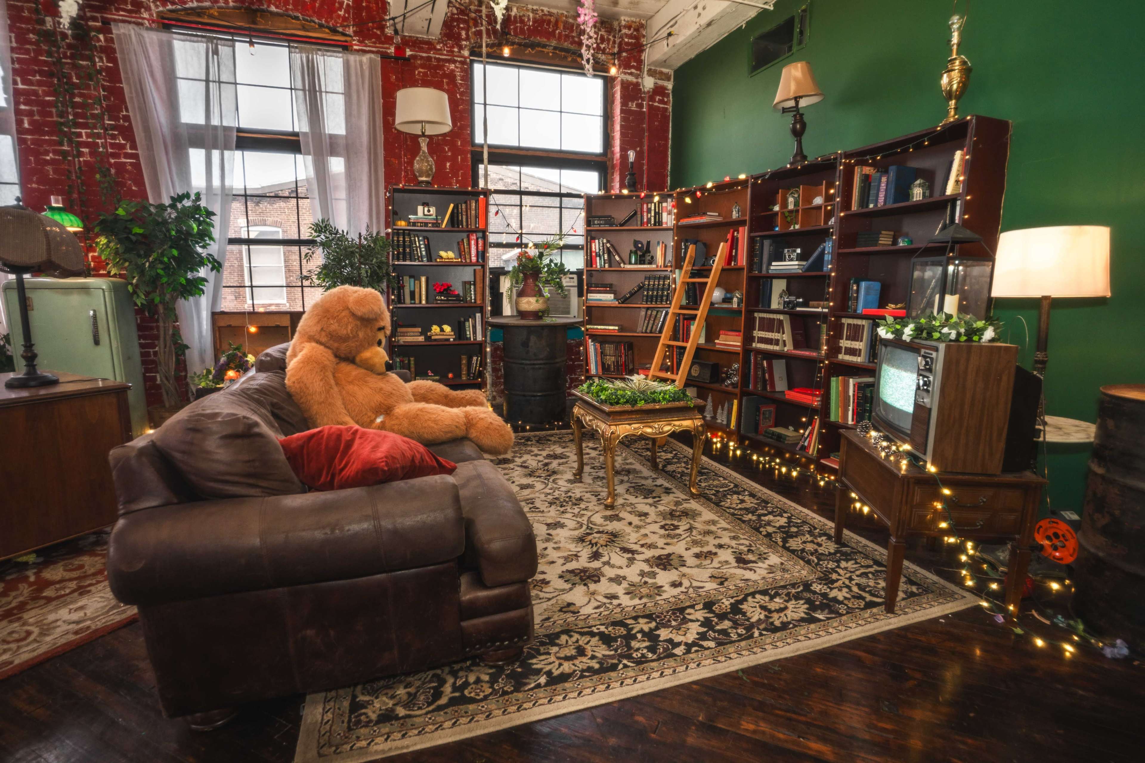 The image shows a cozy, vintage-styled room with a large teddy bear seated on a brown leather couch, surrounded by bookshelves, plants, and soft lighting.