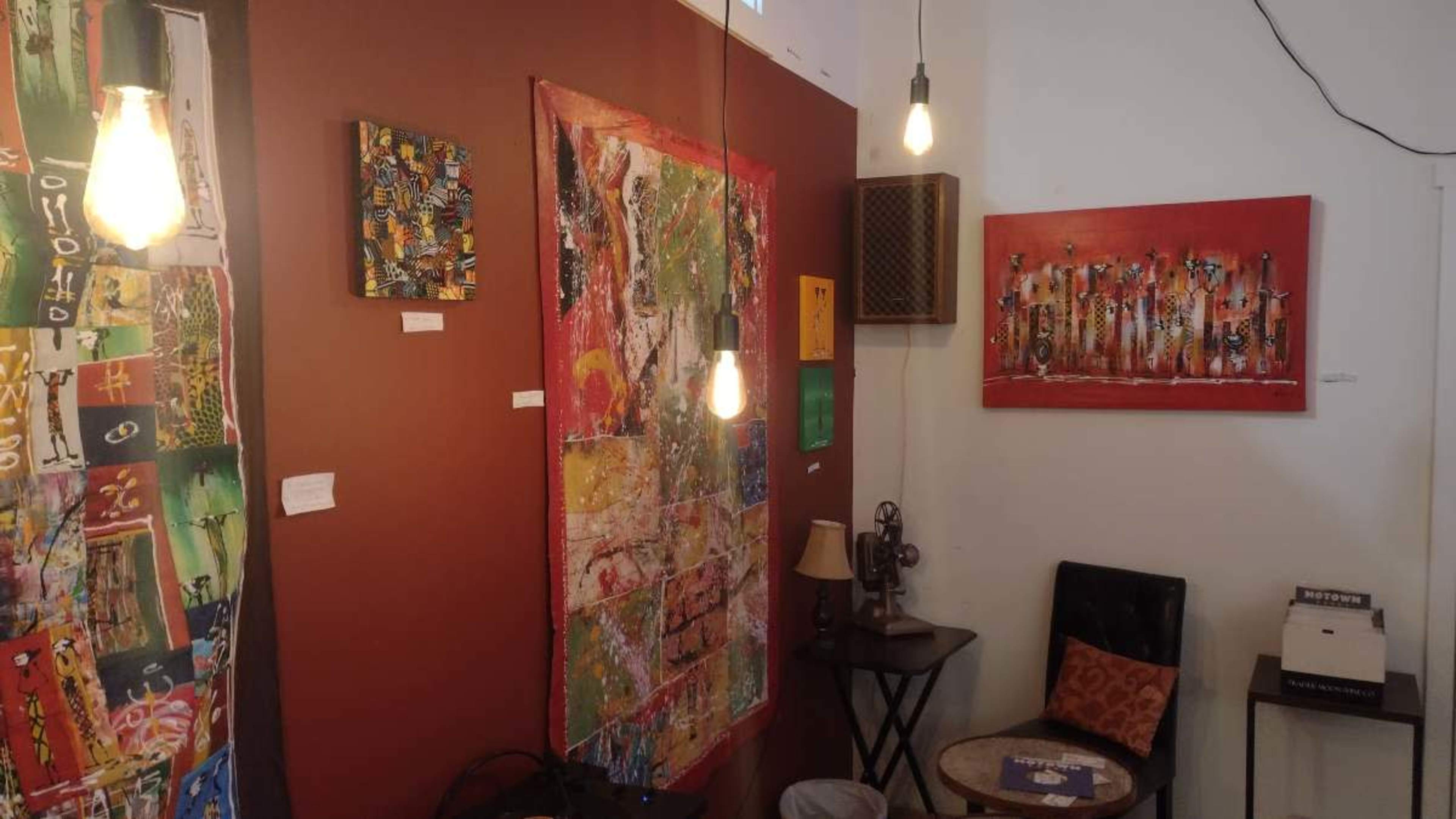 The image shows a cozy art space featuring colorful abstract paintings on the walls, with a small seating area and hanging light bulbs.