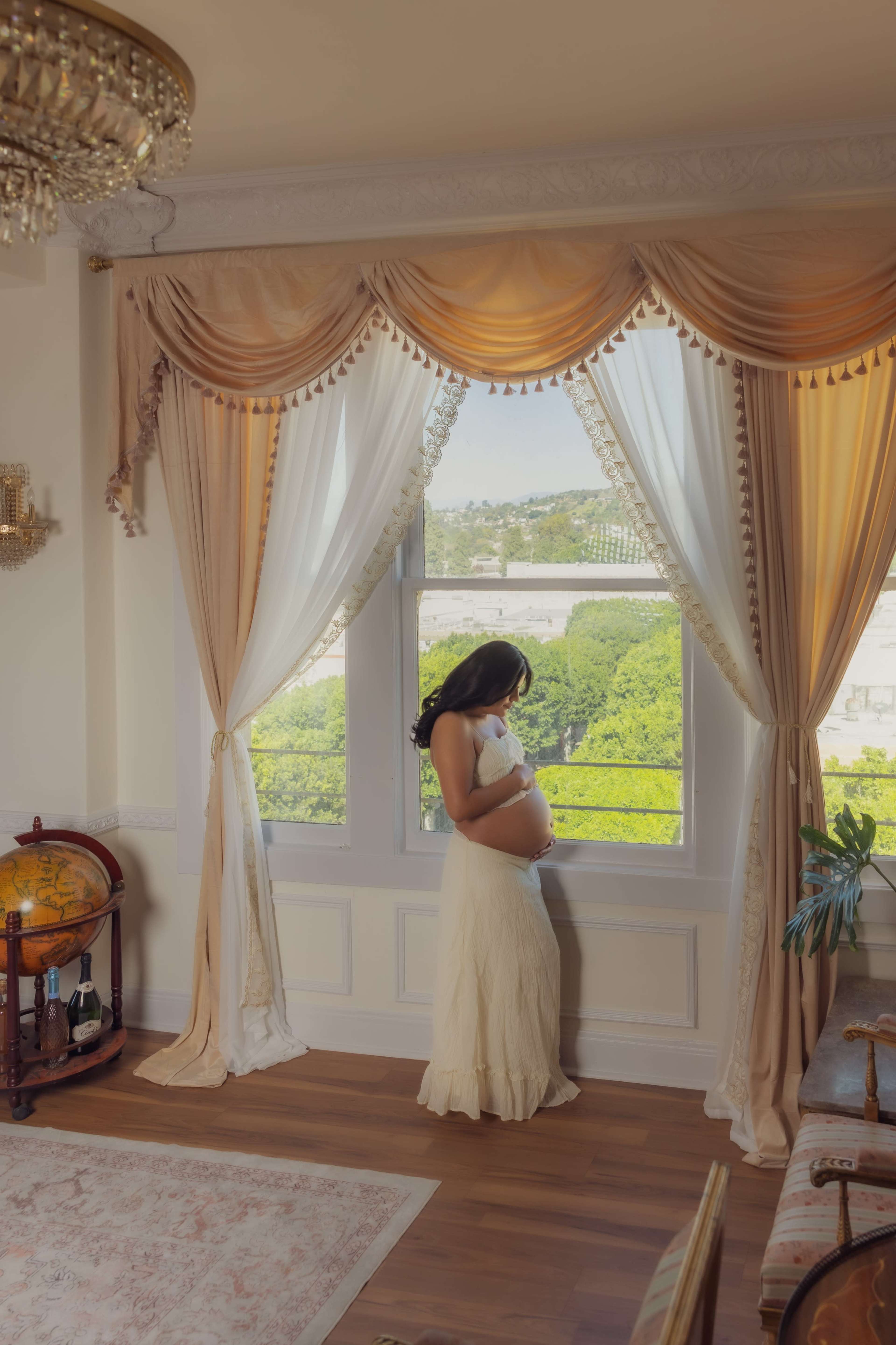 A pregnant woman stands by a large window draped with elegant curtains, gazing out at a lush, green landscape.