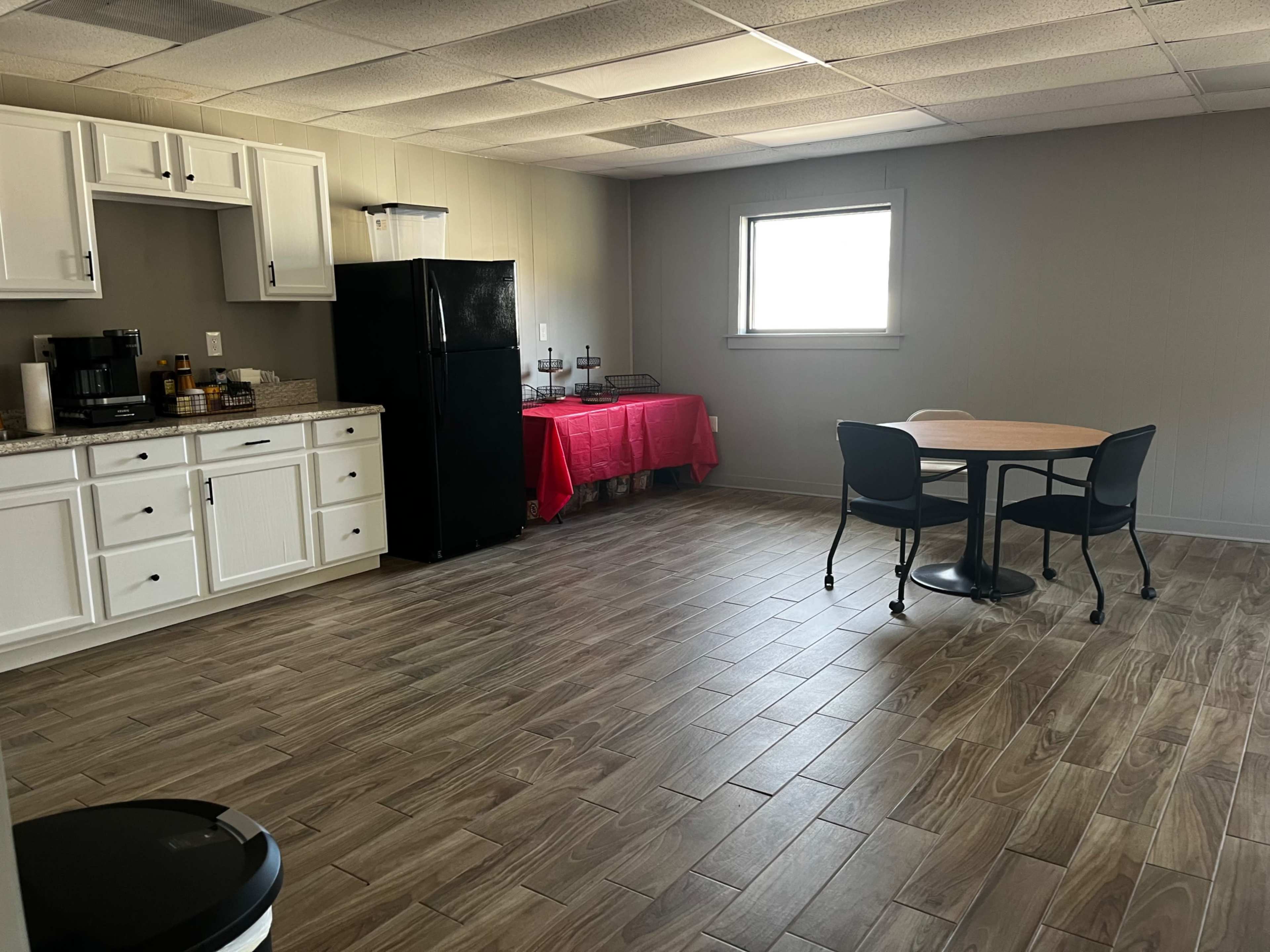 The image shows a kitchen area with white cabinets, a black refrigerator, a countertop, a round table with four chairs, and a red tablecloth-covered table along the back wall.