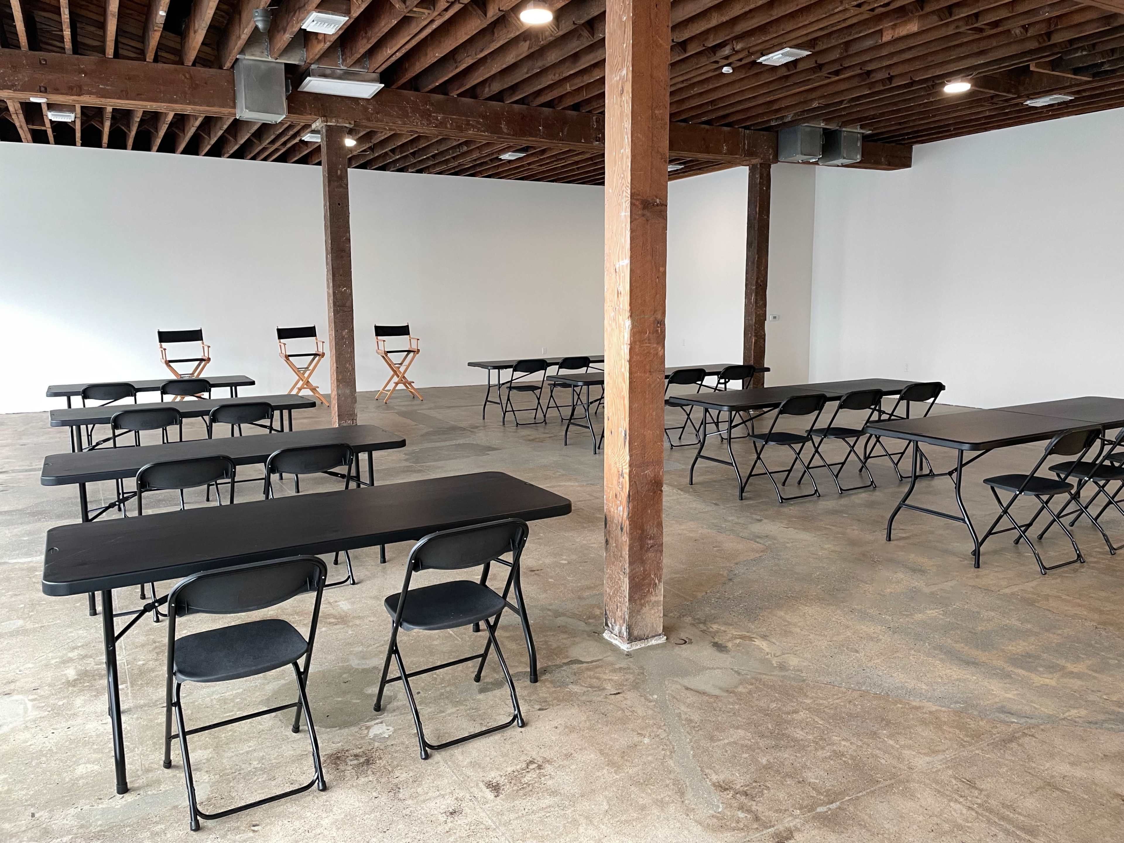 An empty room with wooden beams and a concrete floor features several black folding tables and chairs arranged in a grid pattern, with two director's chairs in the background.