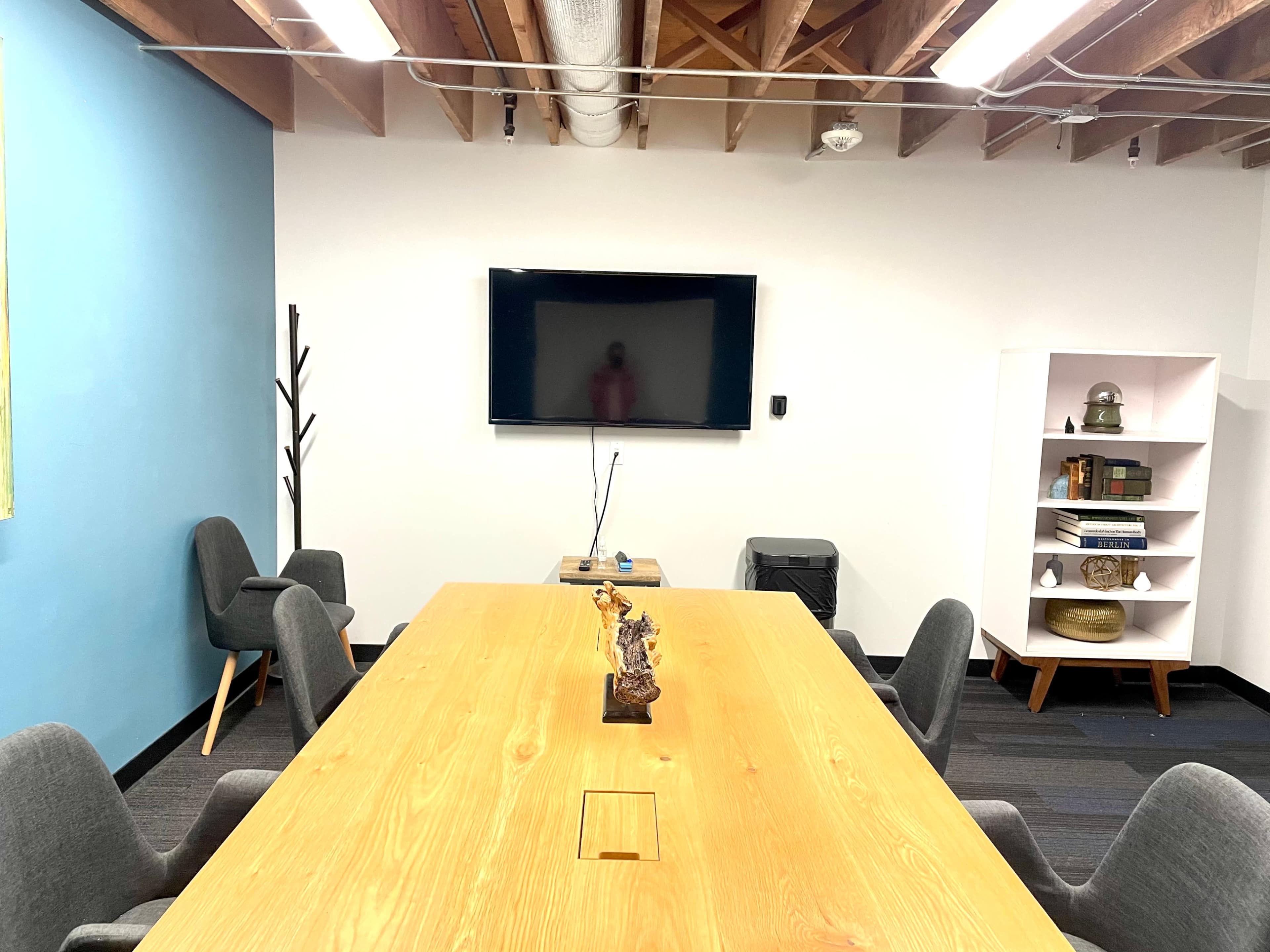 An empty conference room features a large wooden table surrounded by gray chairs, a wall-mounted television, and a white bookshelf against a blue accent wall.