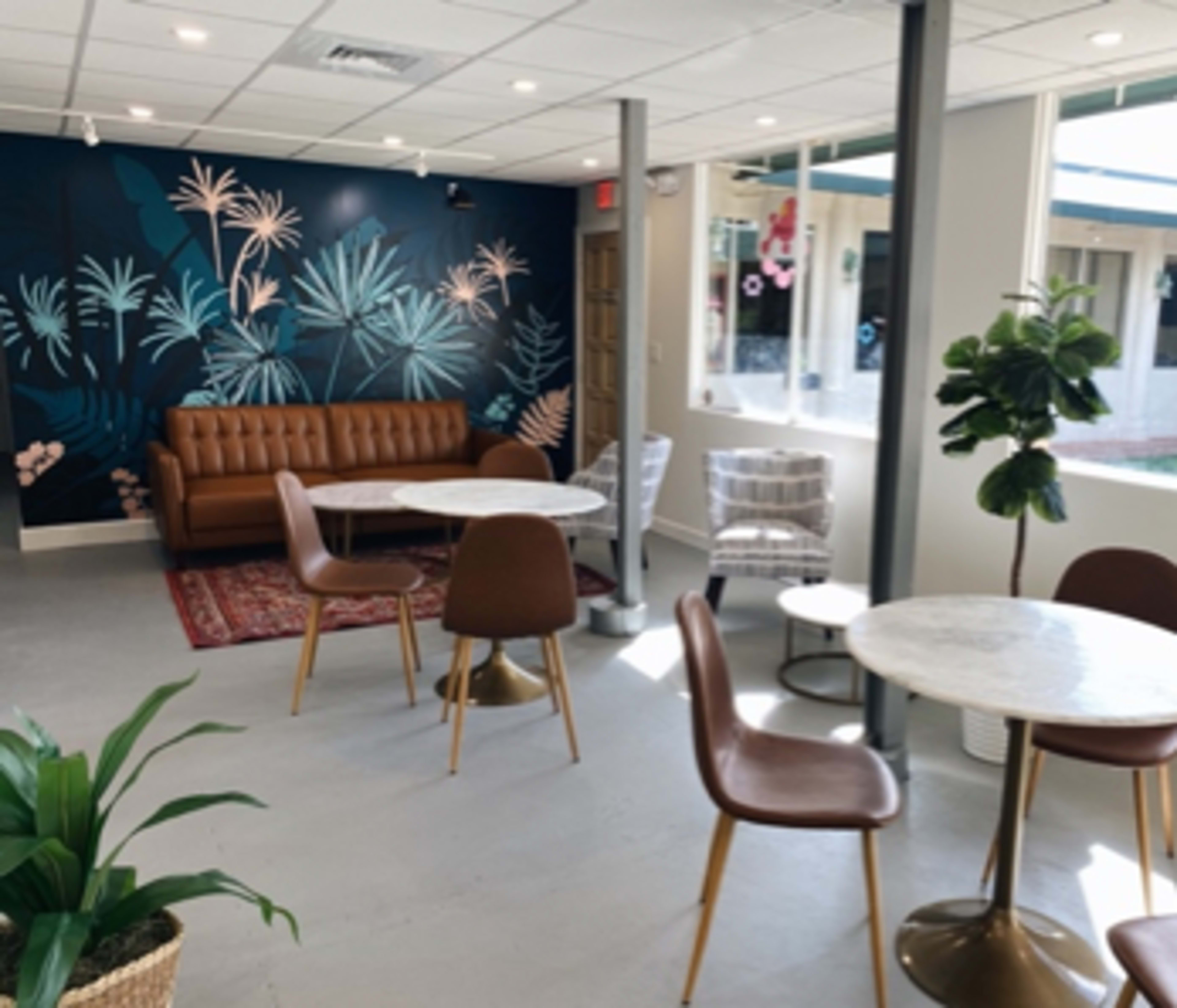 A modern café interior with a brown leather couch, light-colored tables, and floral wall art.