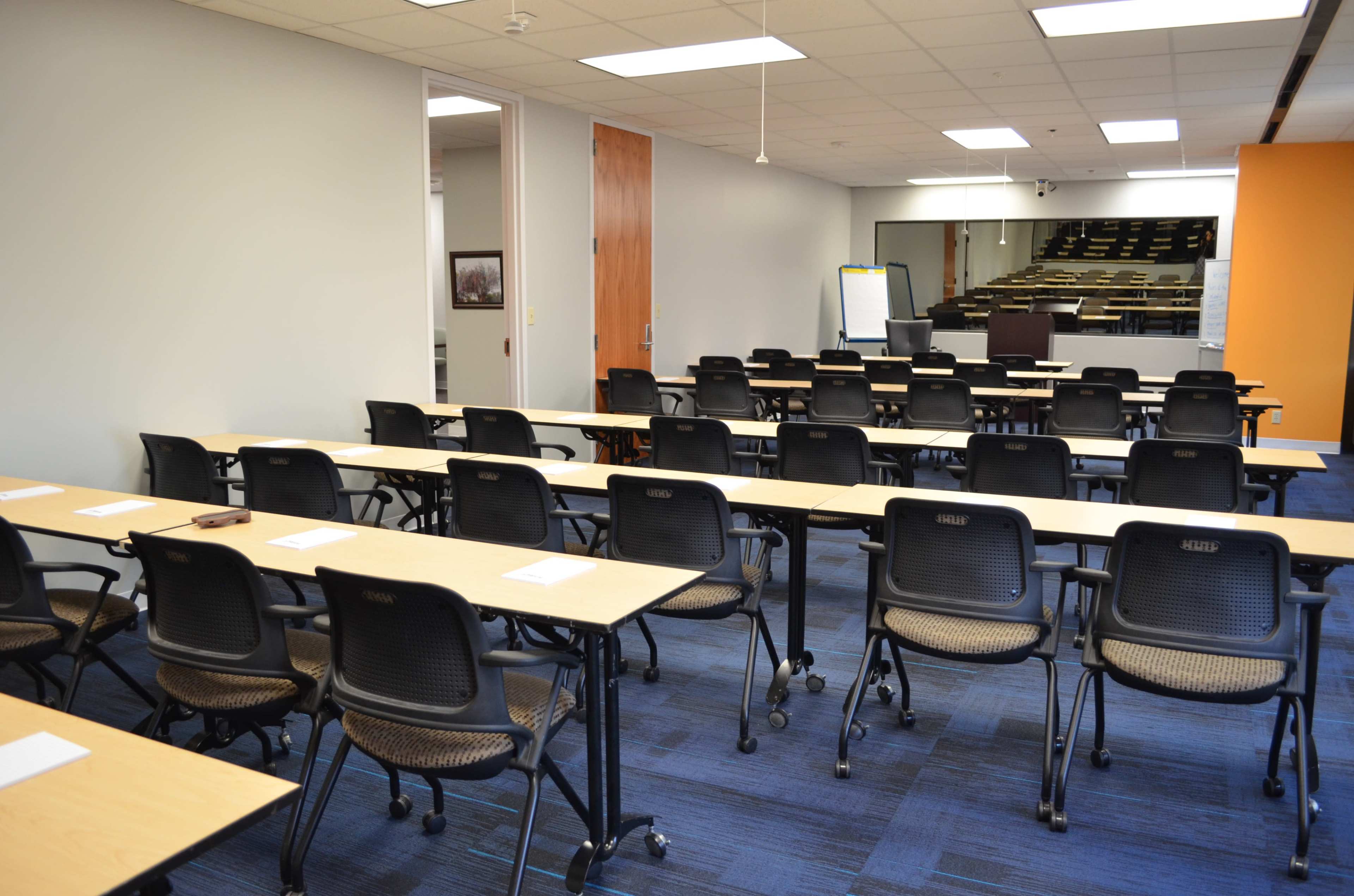 The image shows a classroom setting with multiple rows of tables and chairs arranged for a meeting or training session.
