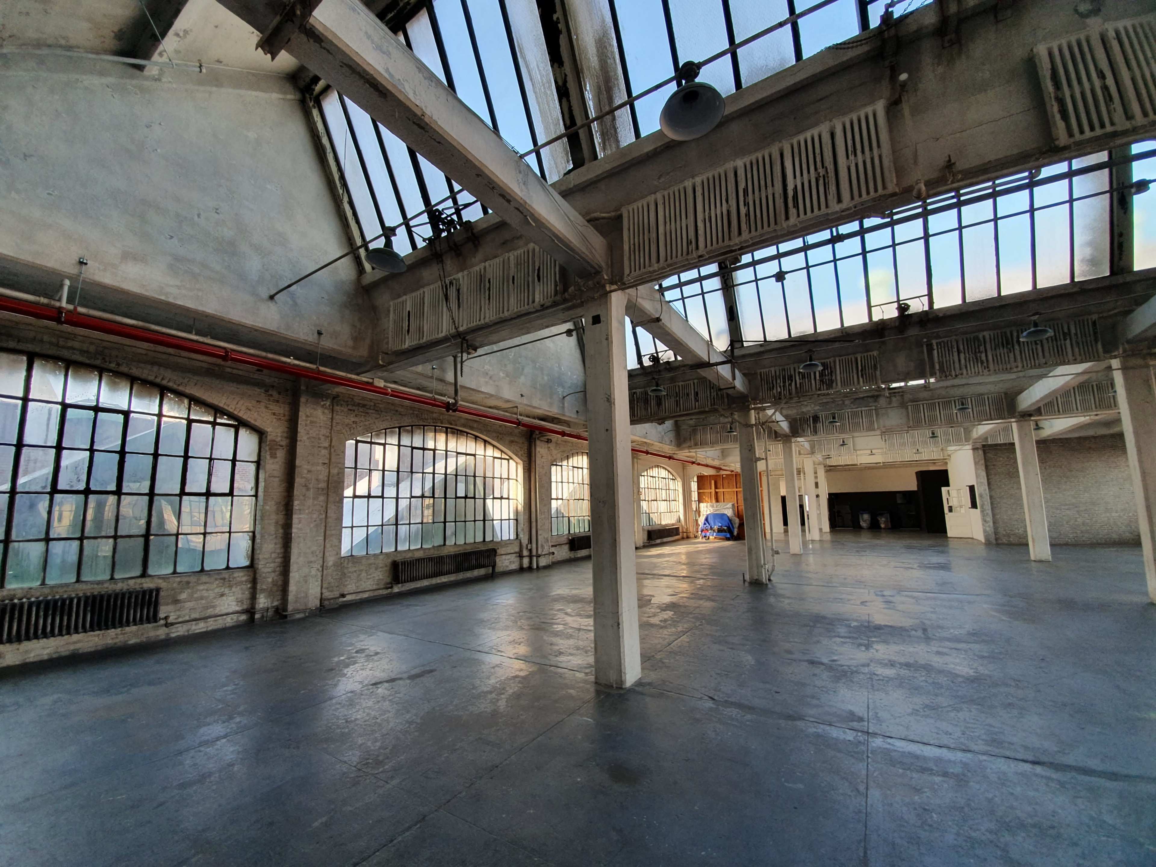 A large, open industrial space with high ceilings, large windows, and a concrete floor.