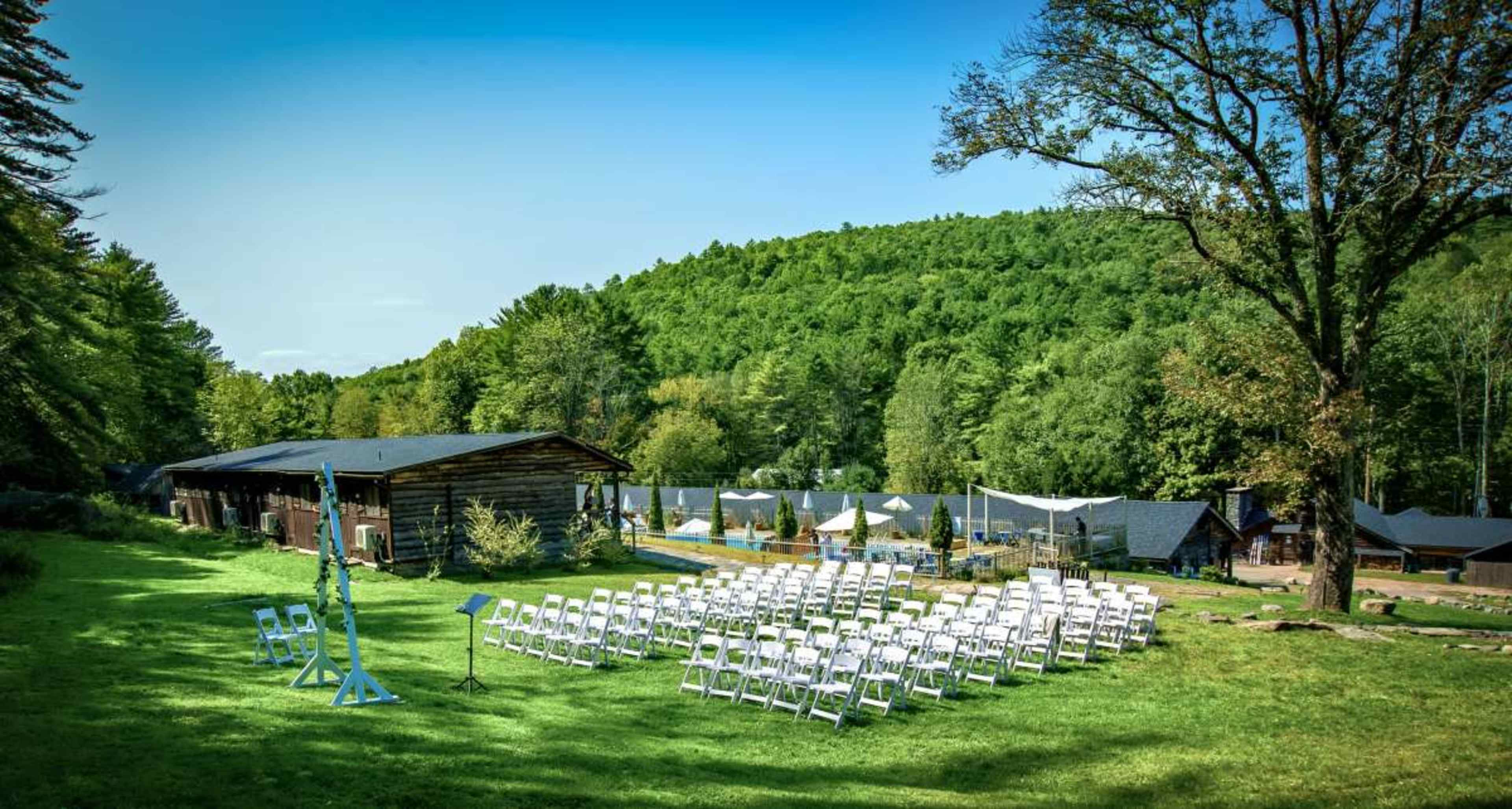 A seating arrangement of white chairs is set up on a grassy area in front of a wooden building, surrounded by trees and hills.